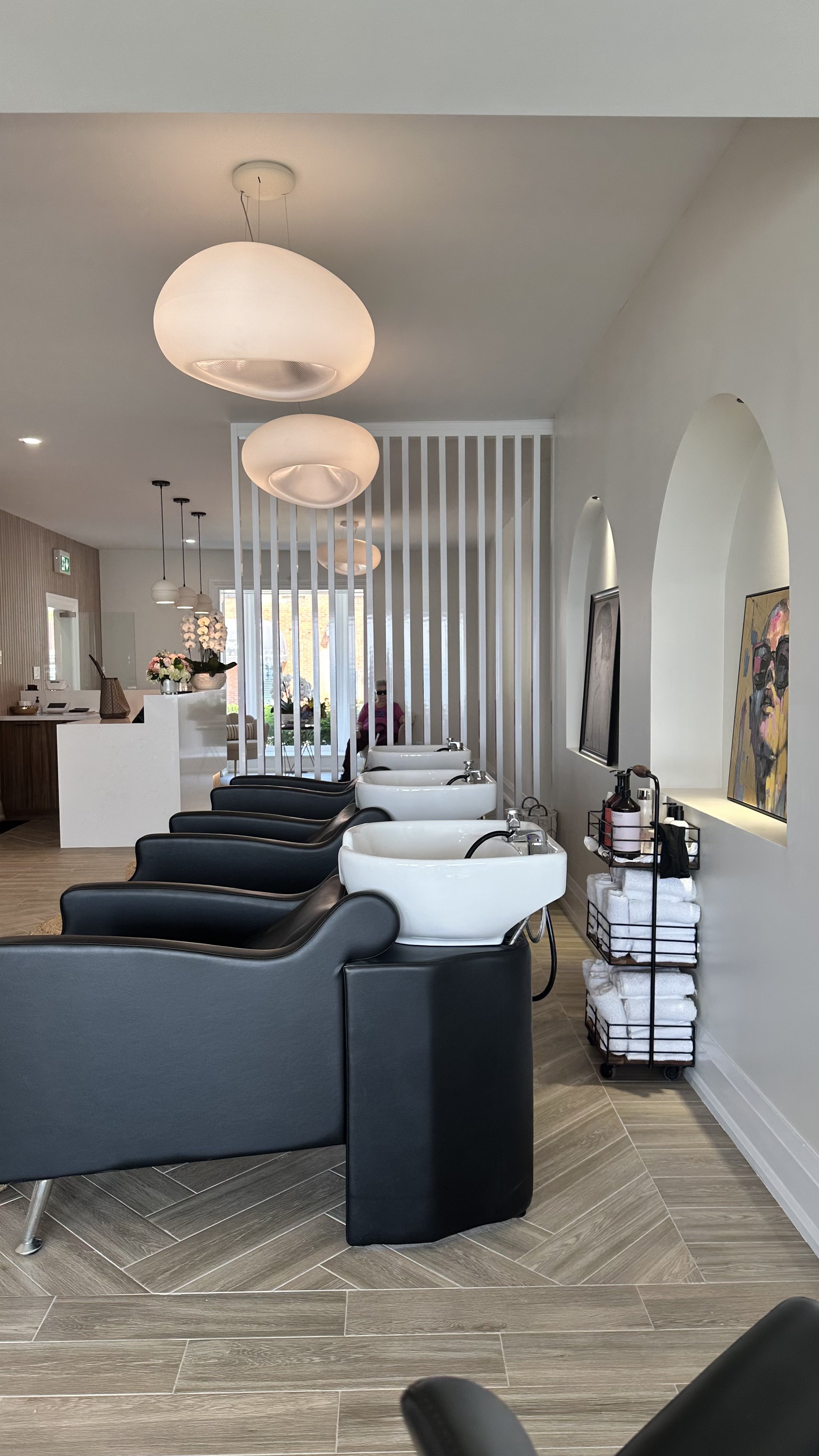 View of a modern hair salon with black and white wash stations, wooden flooring, and contemporary lighting fixtures.