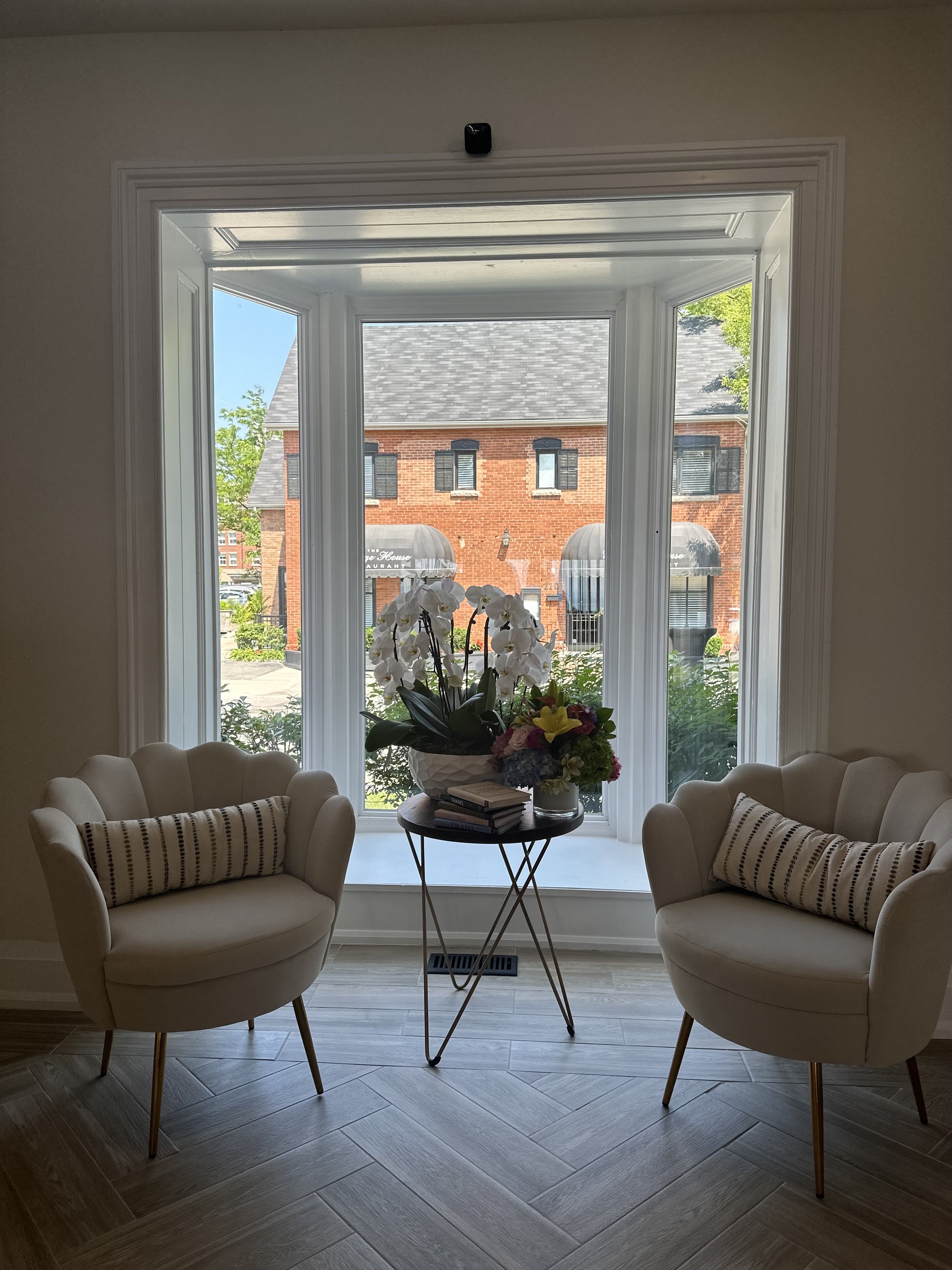 Interior view of a sitting area with two beige armchairs with striped pillows, a round black table with a large potted white orchid and a bouquet of colorful flowers, and a bay window showing a brick building and greenery outside.