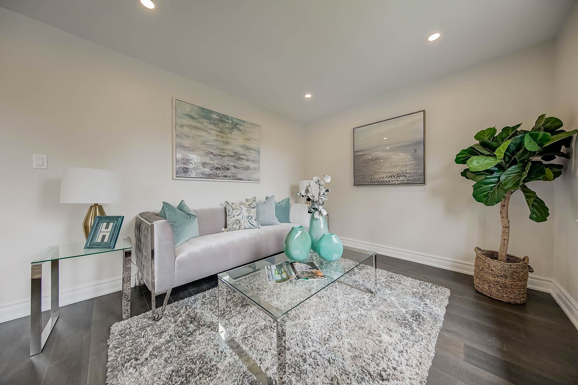 Living room with a modern white sofa, glass coffee table, side table with lamp, potted plant, framed artwork, and decorative vases.