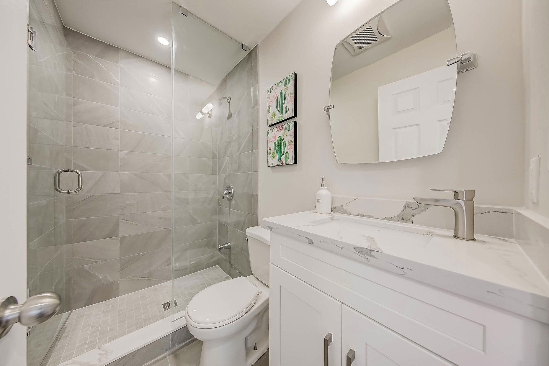 Bathroom with a glass-enclosed shower, white toilet, and white vanity with marble countertop and backsplash. Small wall art with cactus images and a mirror above the sink.