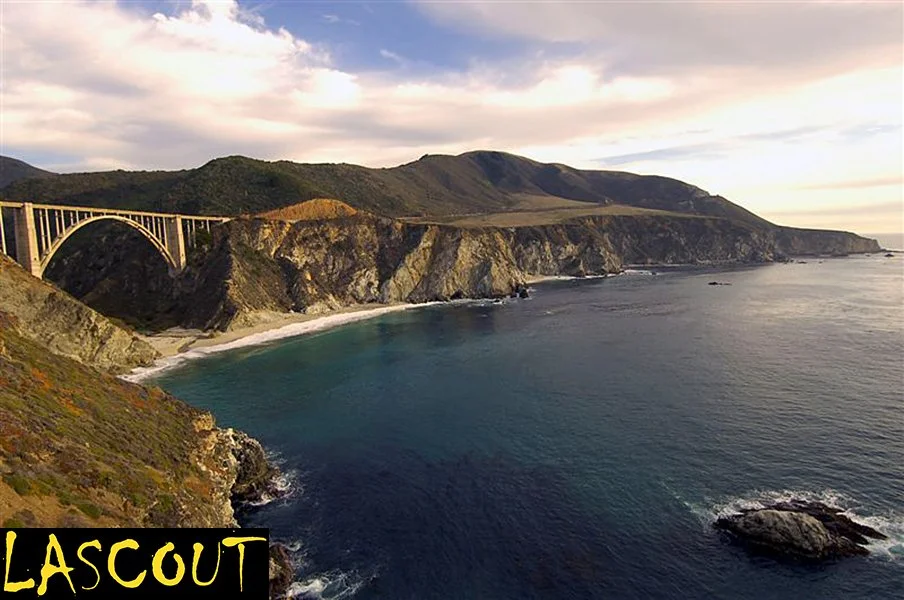 Scenic view of a coastal landscape with cliffs, ocean, and a bridge arching over the hill. The sky has soft clouds and the setting sun.