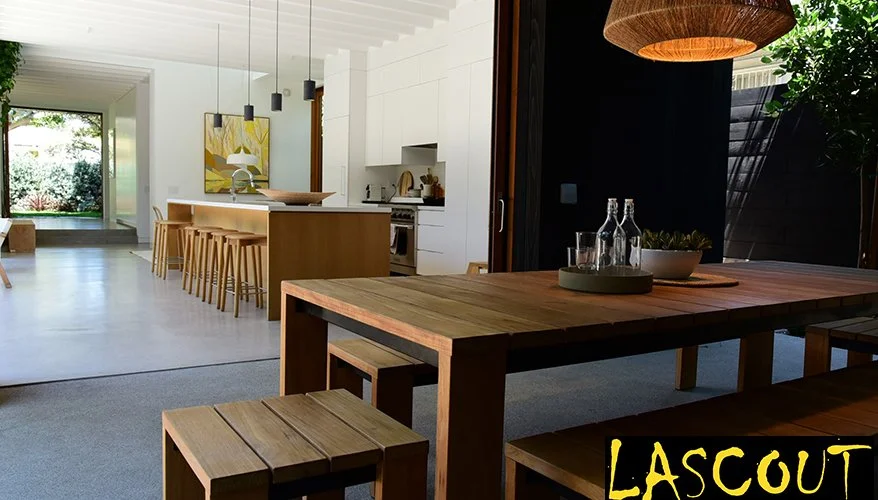 Open-plan kitchen and dining area with wooden furniture, white cabinets, and a large window showing greenery outside.