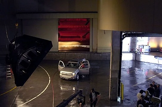 A stage area with a large speaker hanging, a small silver car with open doors, and a backstage area with people and equipment.