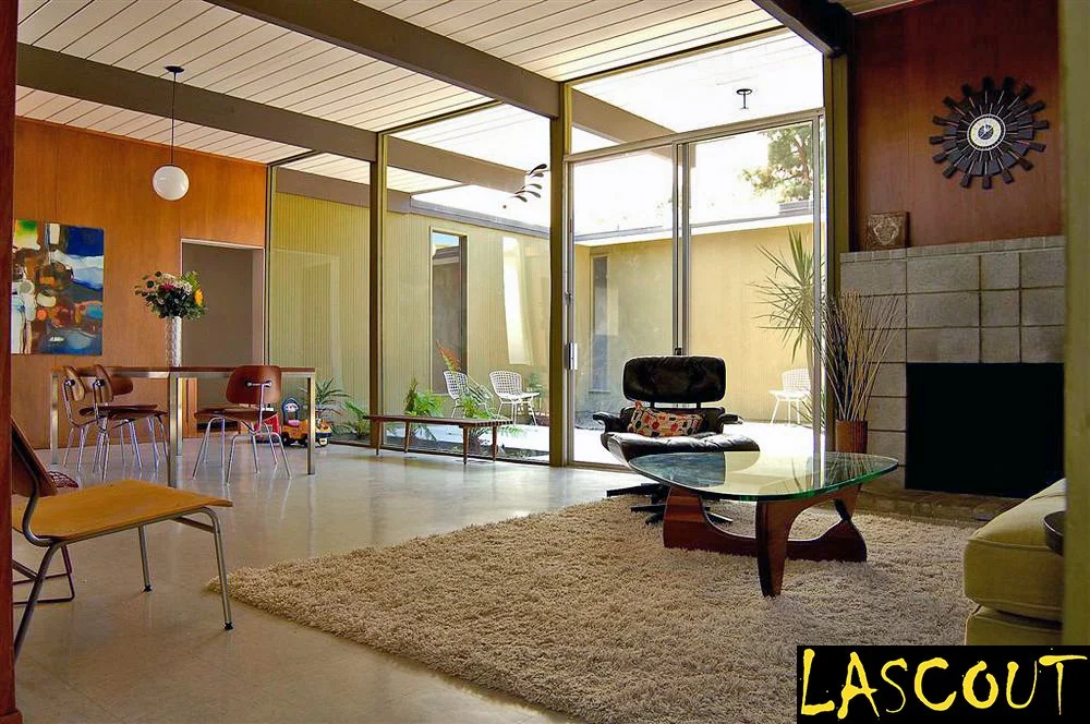 Mid-century modern living room with large glass doors leading to an outdoor patio, featuring a black lounge chair with a matching ottoman, a glass coffee table, a fireplace, and various chairs and plants, in a home with wooden and yellow interior walls.