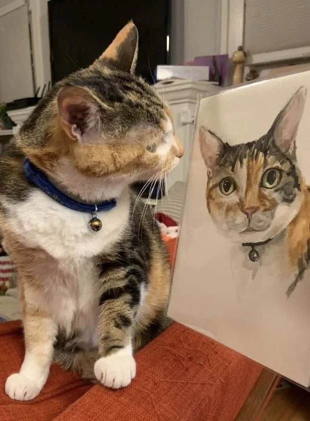 A tabby cat with a blue collar and bell sitting on a table, looking at a watercolor painting of itself.