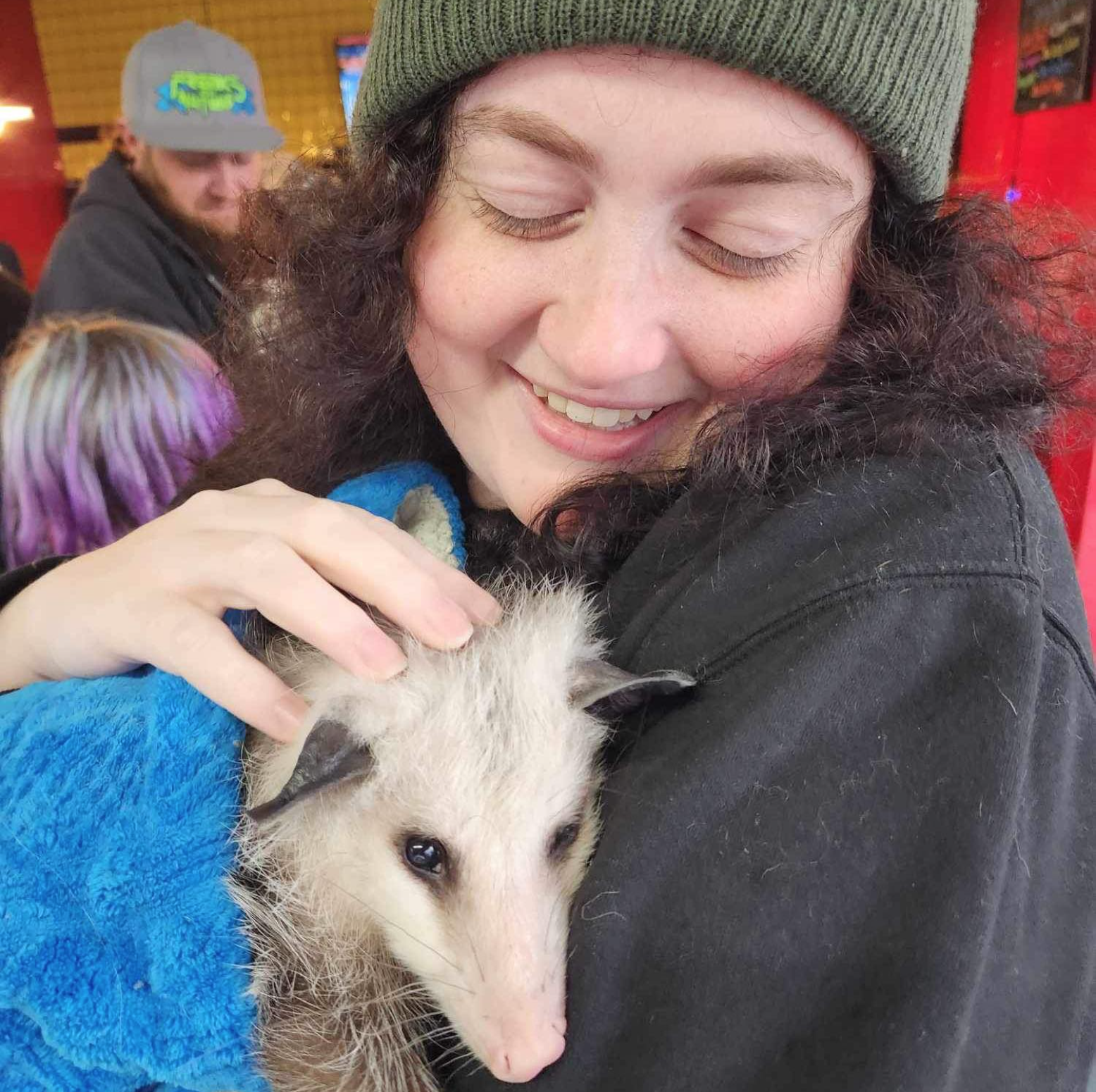 A young woman with curly dark hair, wearing a green beanie, hugging a possum with a content smile.