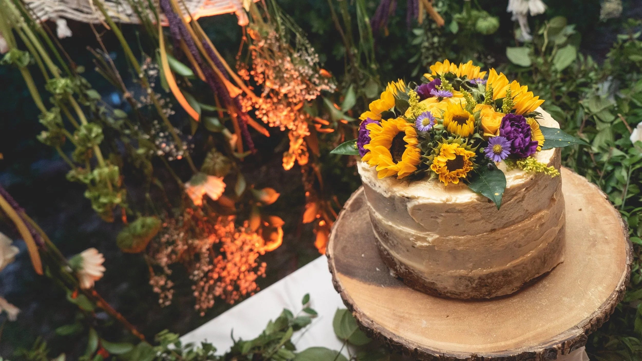 A rustic cake with brown frosting topped with yellow, purple, and white flowers, placed on a wooden slab. The background includes green foliage and warm lighting.