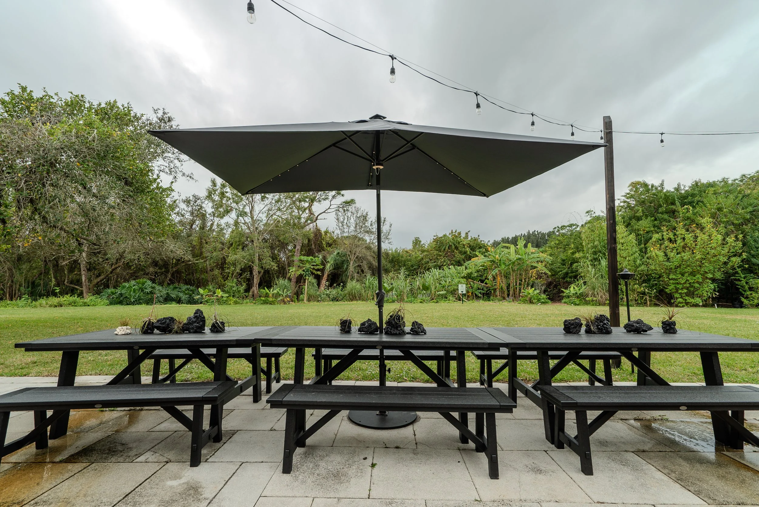 Outdoor patio with black benches and tables, a large black umbrella, black rocks, and small plants, with green trees and cloudy sky in the background.
