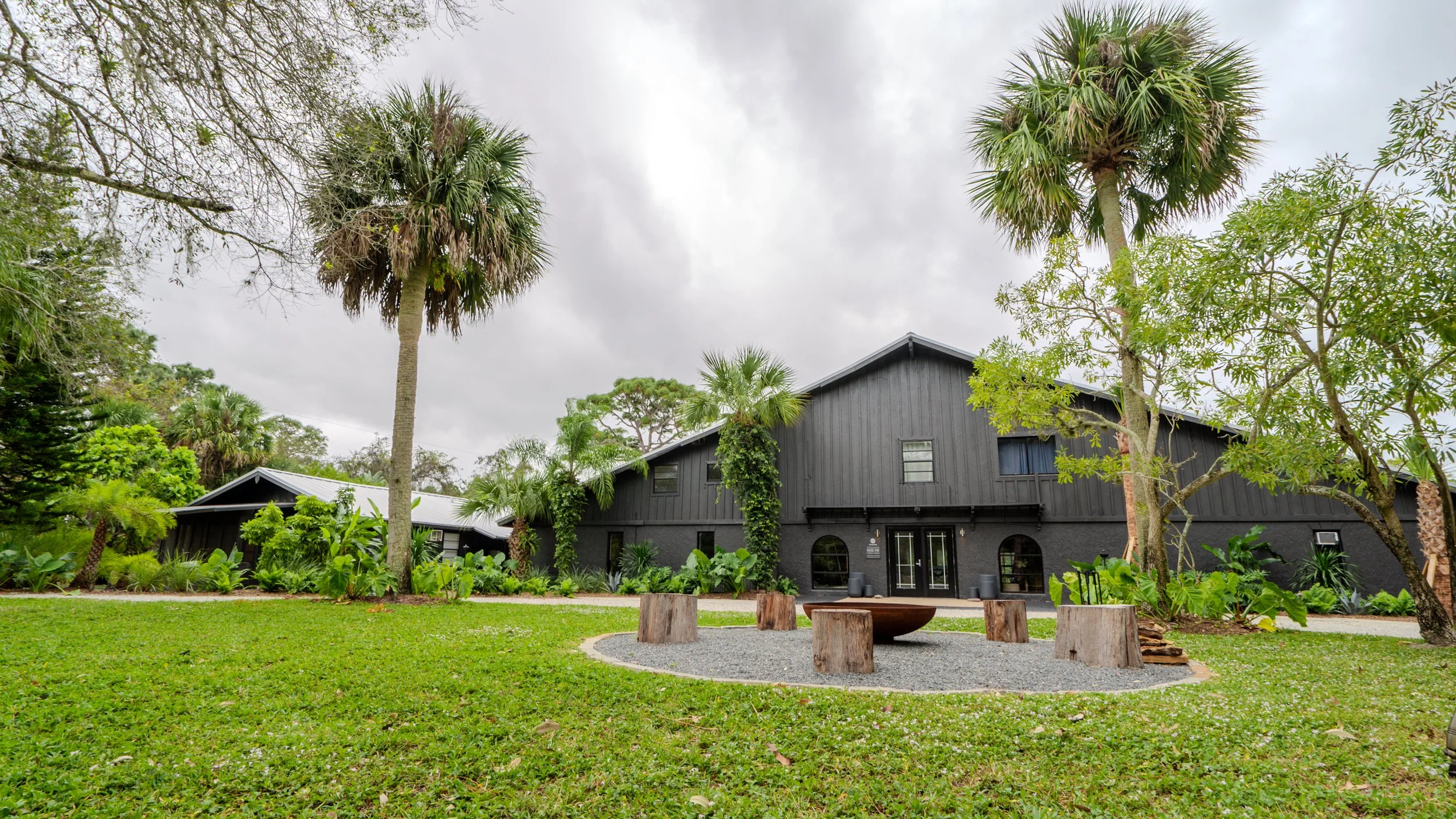A large black house with a metal roof, surrounded by palm trees and greenery, situated in a garden with a circular gravel area featuring wooden stumps and a fire pit.