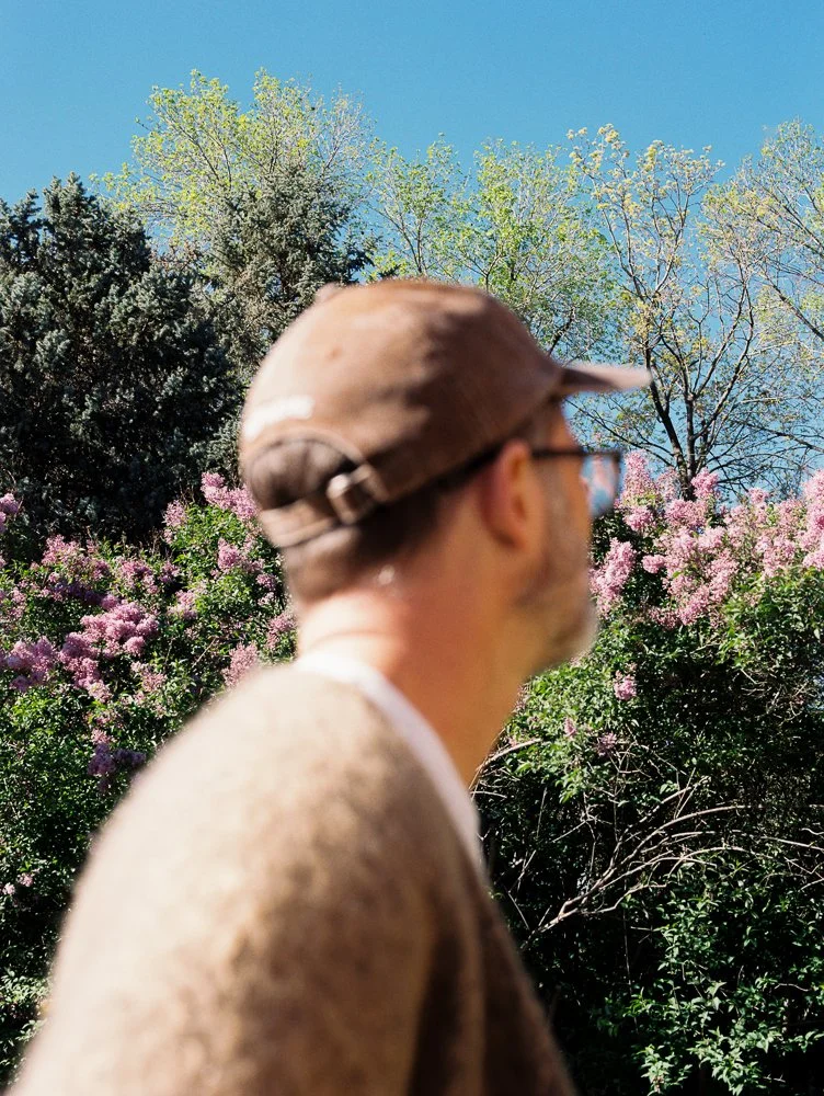 man wearing brown supreme pigment hat in front of lilacs and trees