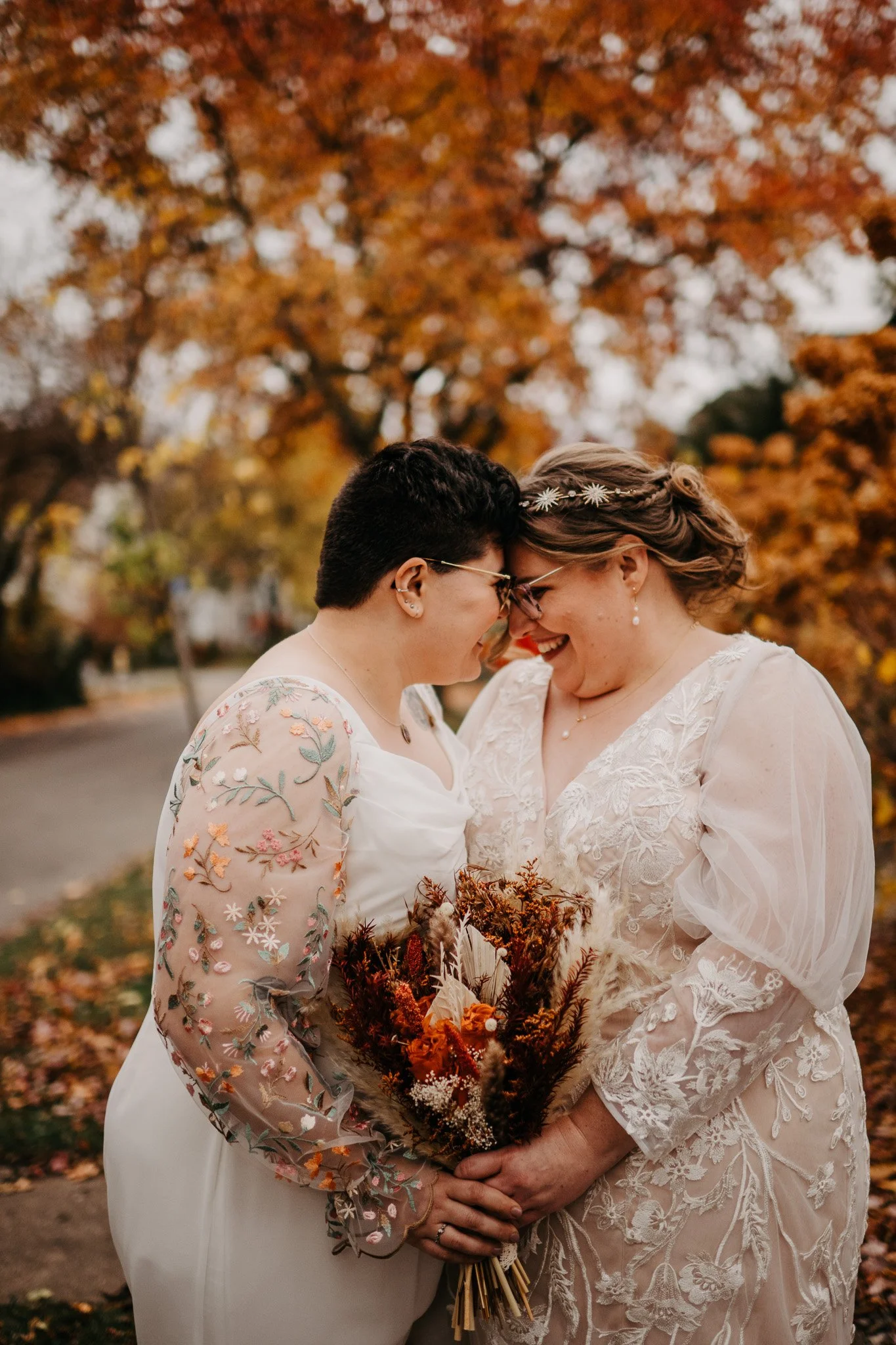 Two brides in embroidered and lace gowns share a joyful forehead touch beneath vibrant fall foliage during their autumn wedding celebration in Rochester, NY.