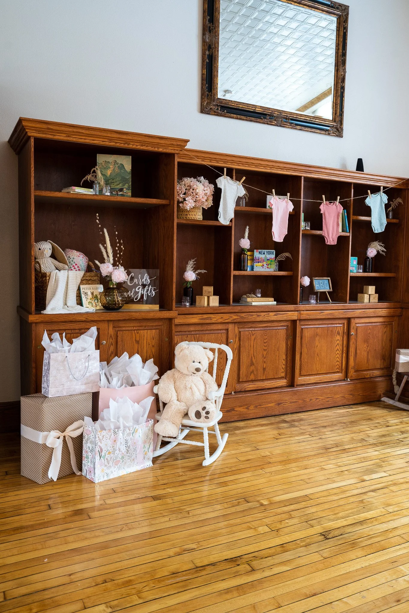 Baby shower gift display in front of wooden built-in shelves, decorated with pink and blue onesies, children’s books, and a rocking chair with a teddy bear.