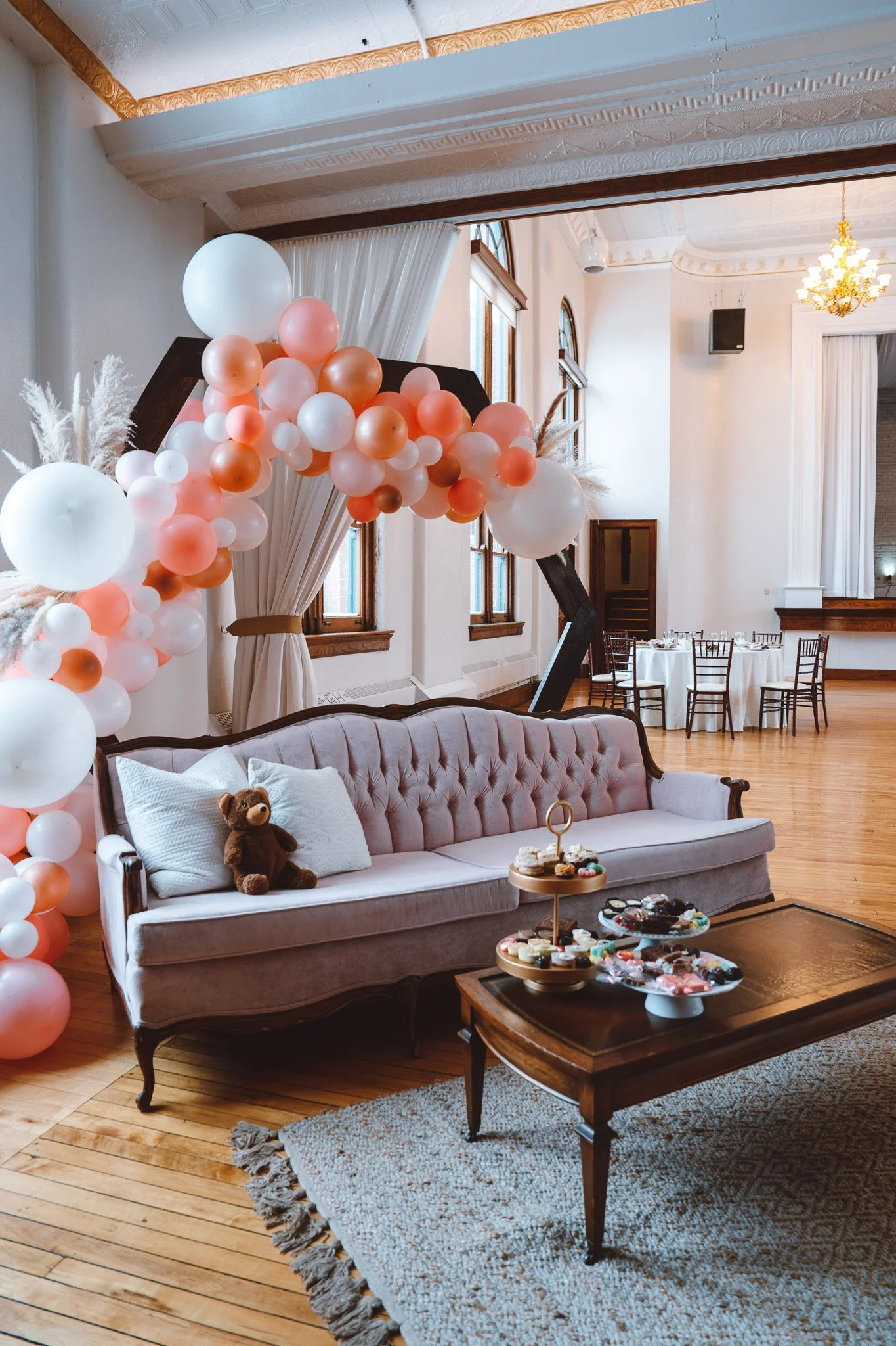Pink, peach, and white balloon garland arching over a mauve velvet loveseat with dessert trays on a coffee table, set beneath The Historic German House’s tall windows and tin ceiling.