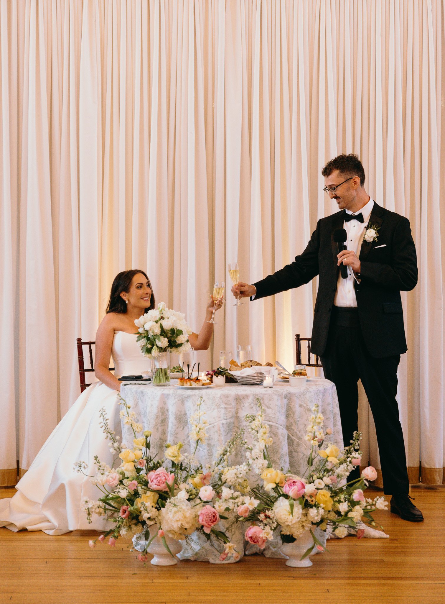 Bride and groom clinking champagne glasses during a sweetheart table toast, surrounded by lush floral arrangements at The Historic German Houyse