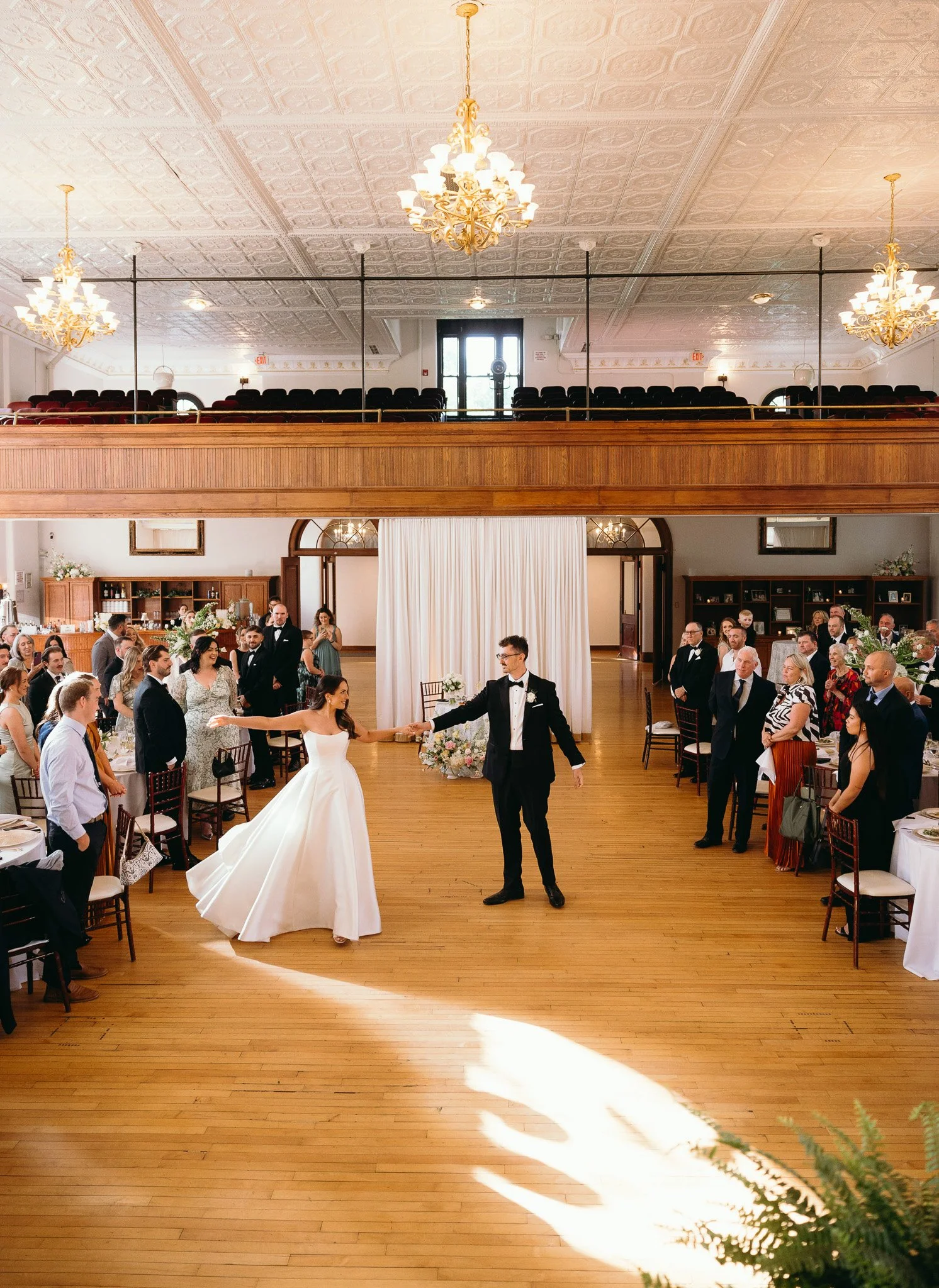 Newly married couple holding hands during their first dance at the reception, surrounded by cheering guests and vintage chandeliers.
