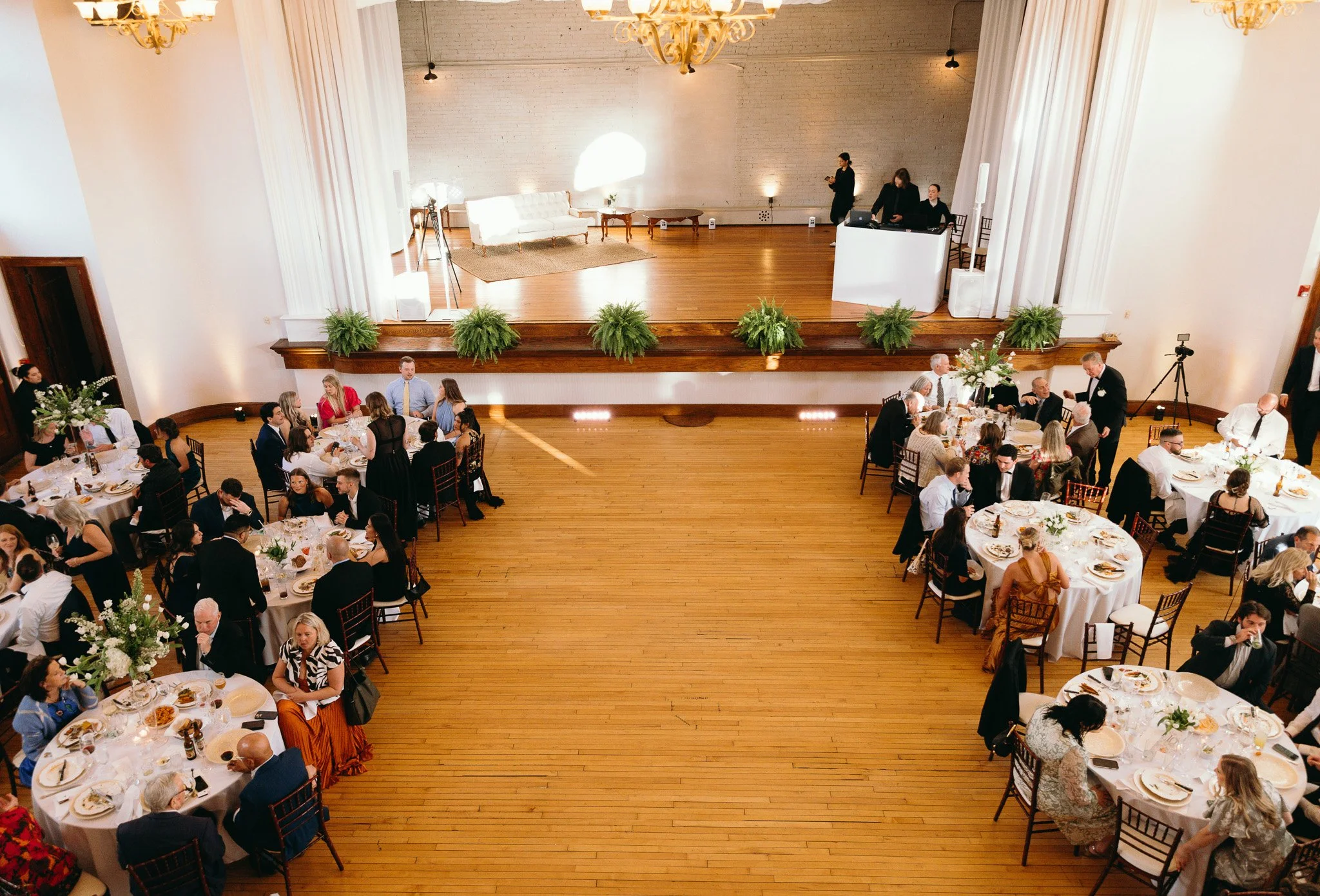Overhead view of an event inside The Historic German House, with guests seated at round tables surrounding a vintage stage with white draping and greenery.