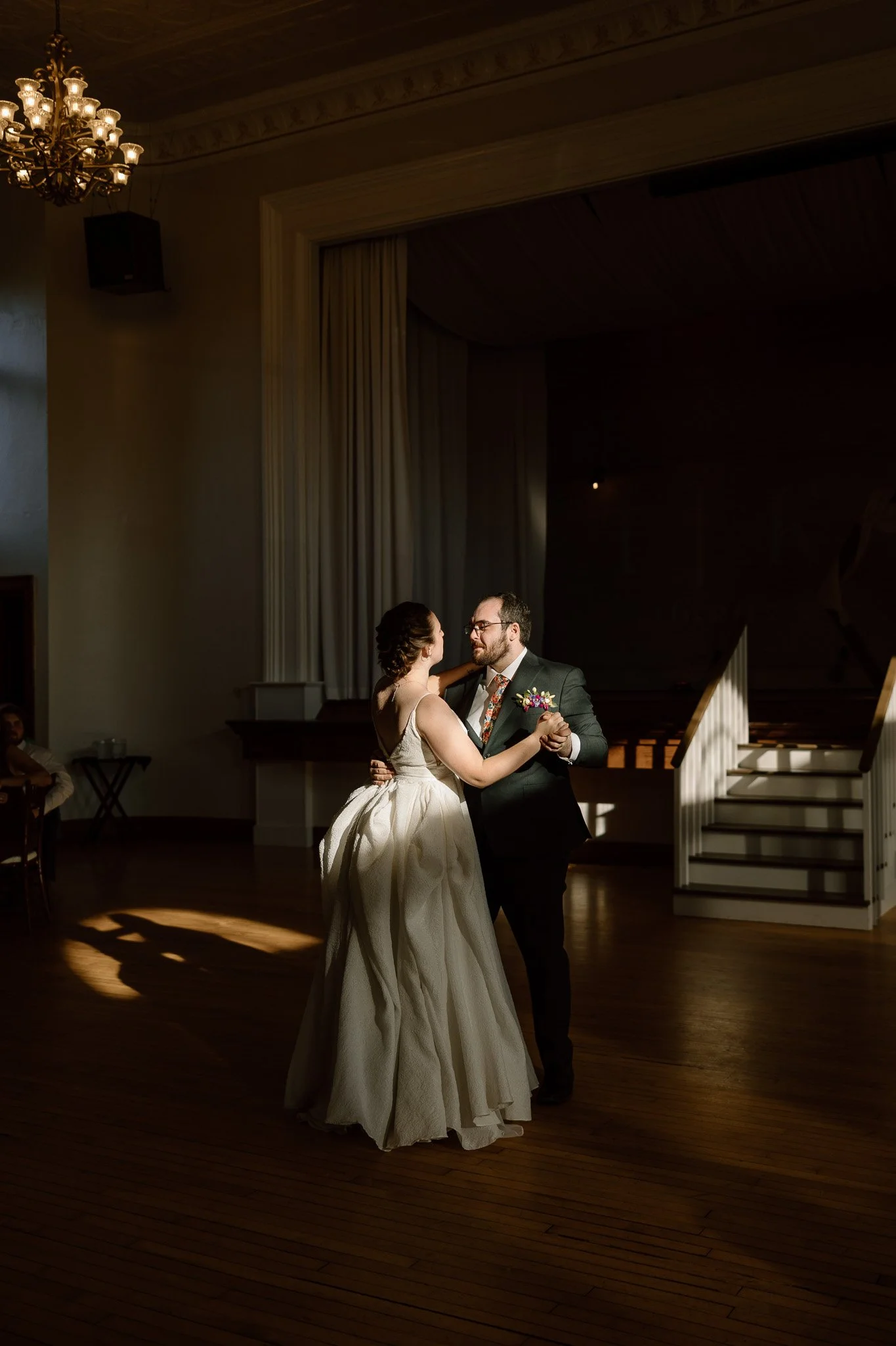 Bride and groom sharing their first dance under a chandelier, illuminated by a dramatic beam of light in a vintage ballroom with wood flooring.
