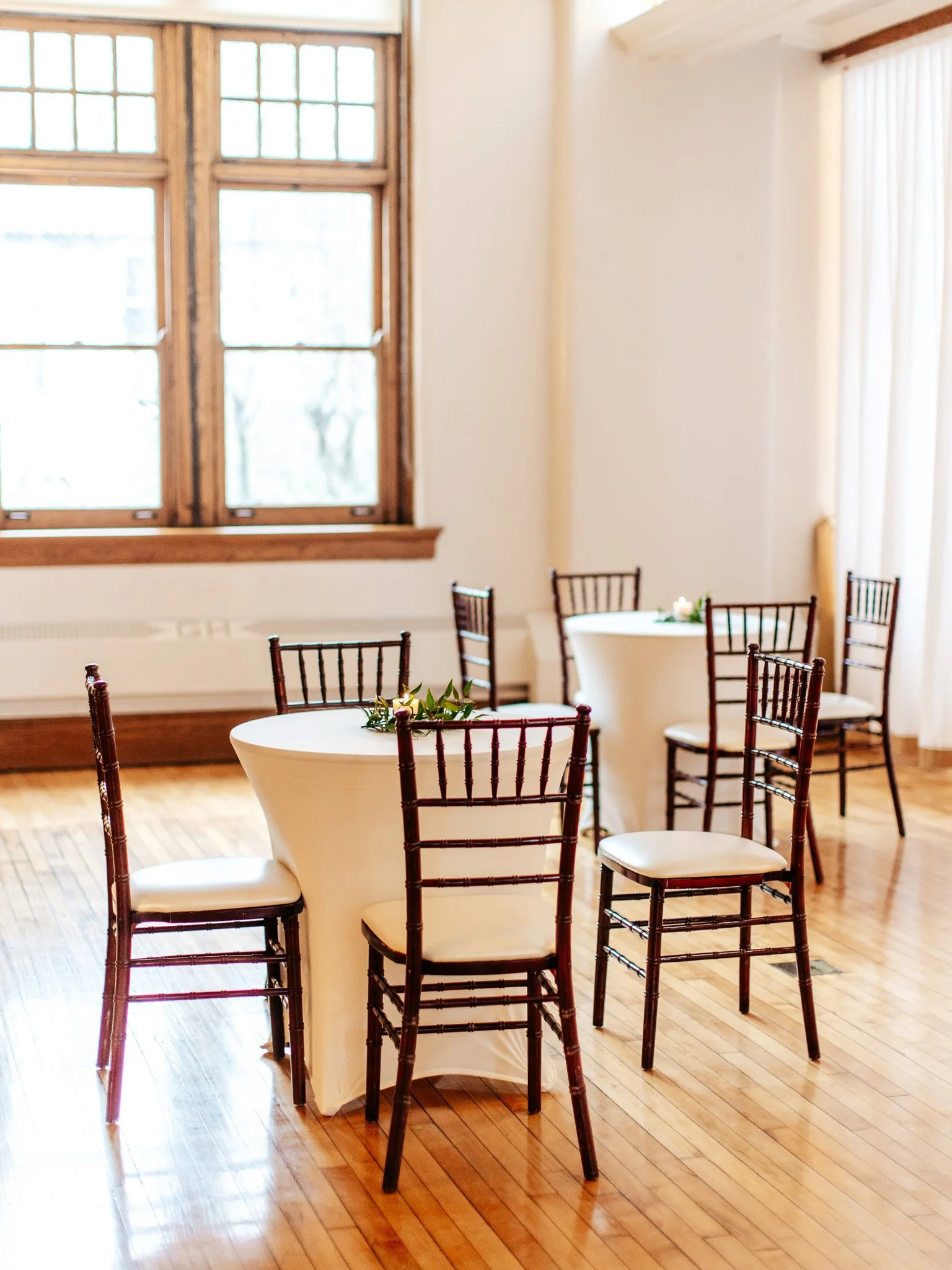 Natural light filtering through tall windows onto intimate cocktail tables with white linens and dark wooden Chiavari chairs at The Historic German House.