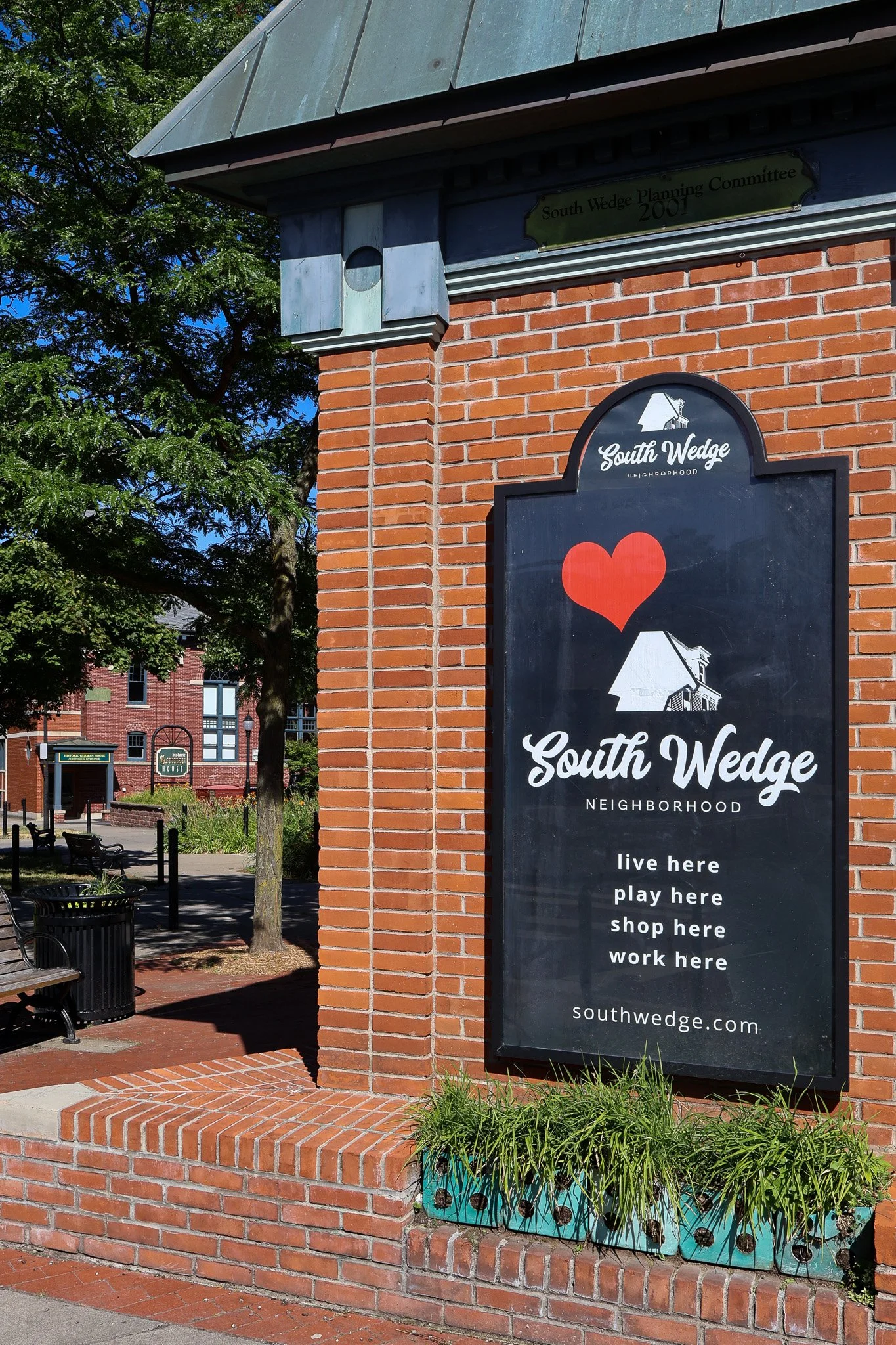 South Wedge Neighborhood sign in the center of the neighborhood, surrounded by brick, plants, benches, and in the distance is The Historic German House, in Rochester NY