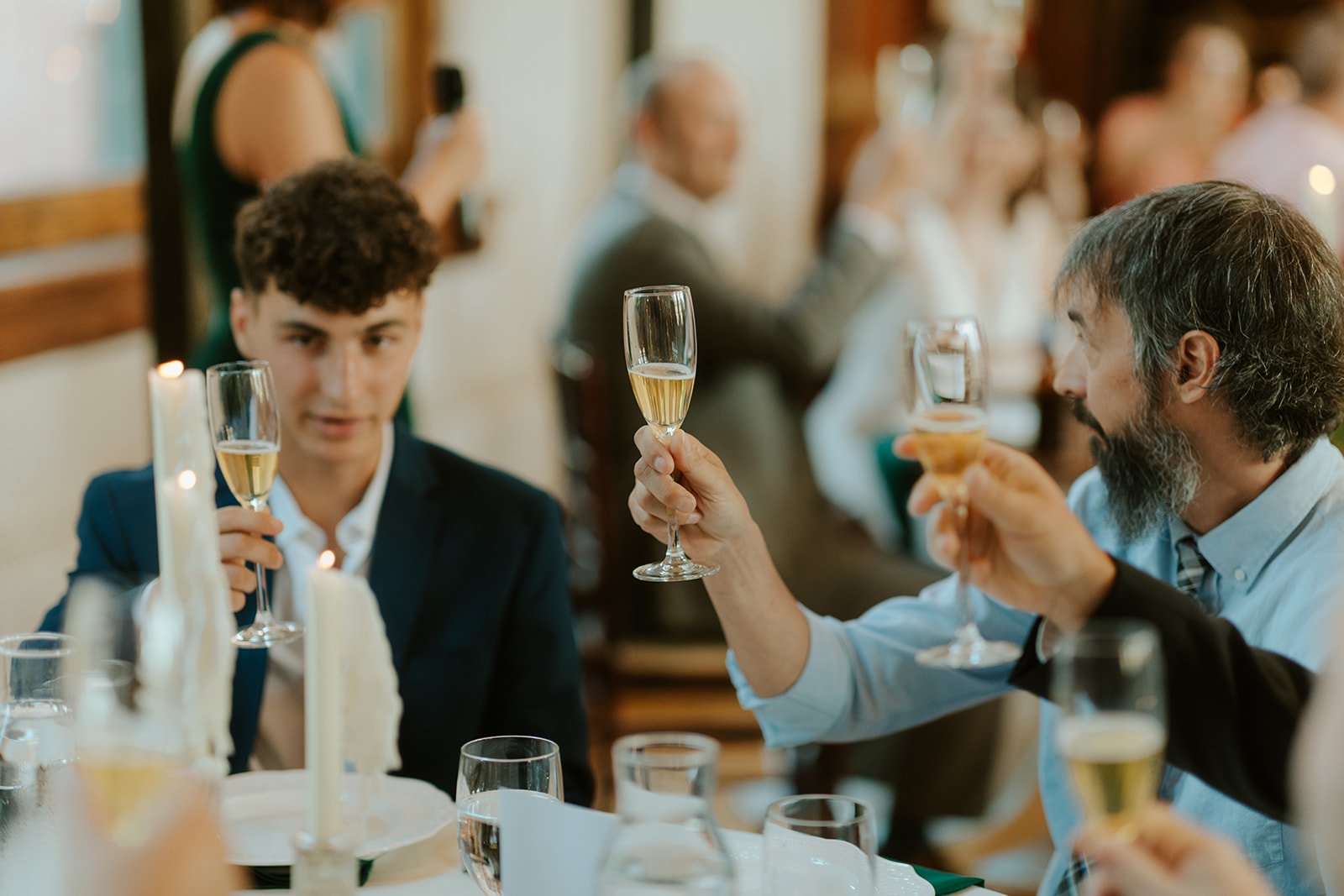 Close-up of guests raising champagne glasses during a wedding toast, with warm candlelight and blurred celebration in the background.