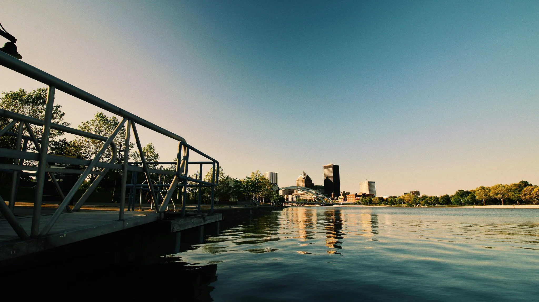 Scenic view of downtown Rochester skyline taken from a metal dock on the Genesee River during golden hour, with reflections on calm water.