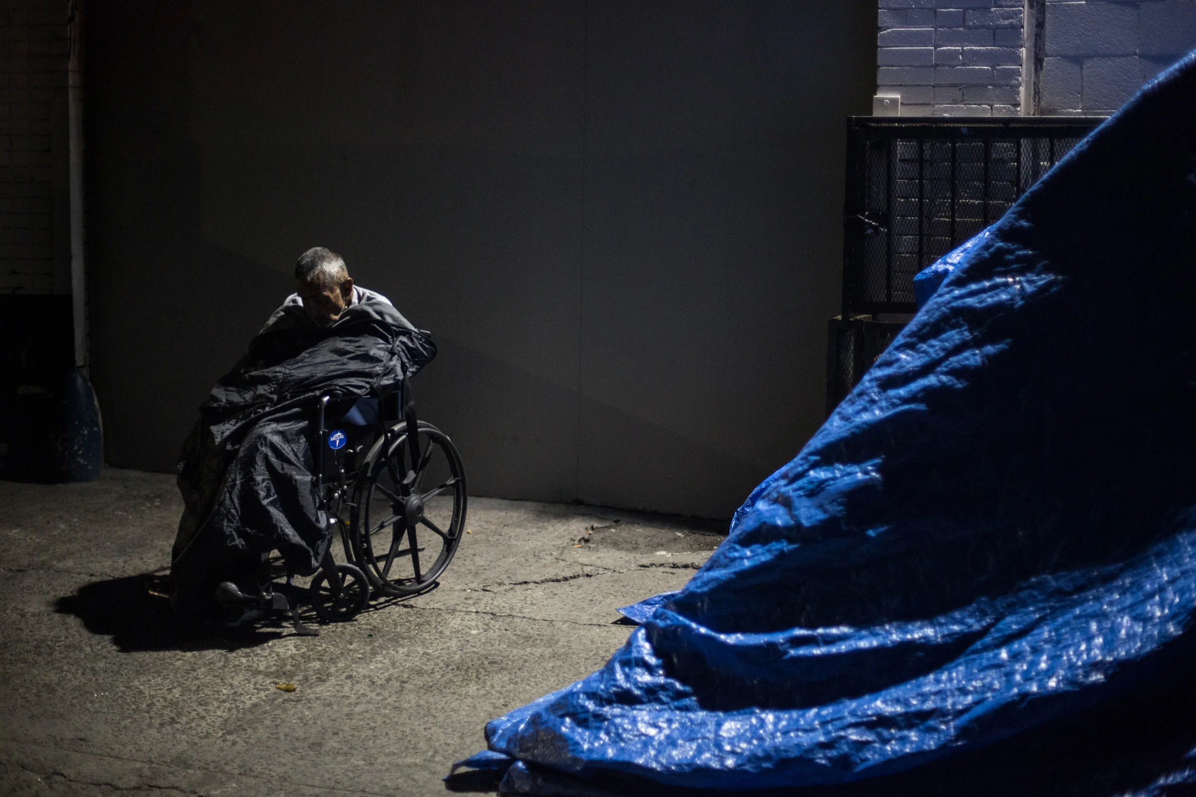 Beneath a street light on Skid Row, an elderly man dozes off under a sleeping bag in Los Angeles, California. Next to him is a large tent made of blue tarp and found material. Apr. 2, 2017 (Staff Photographer Zin Chiang)