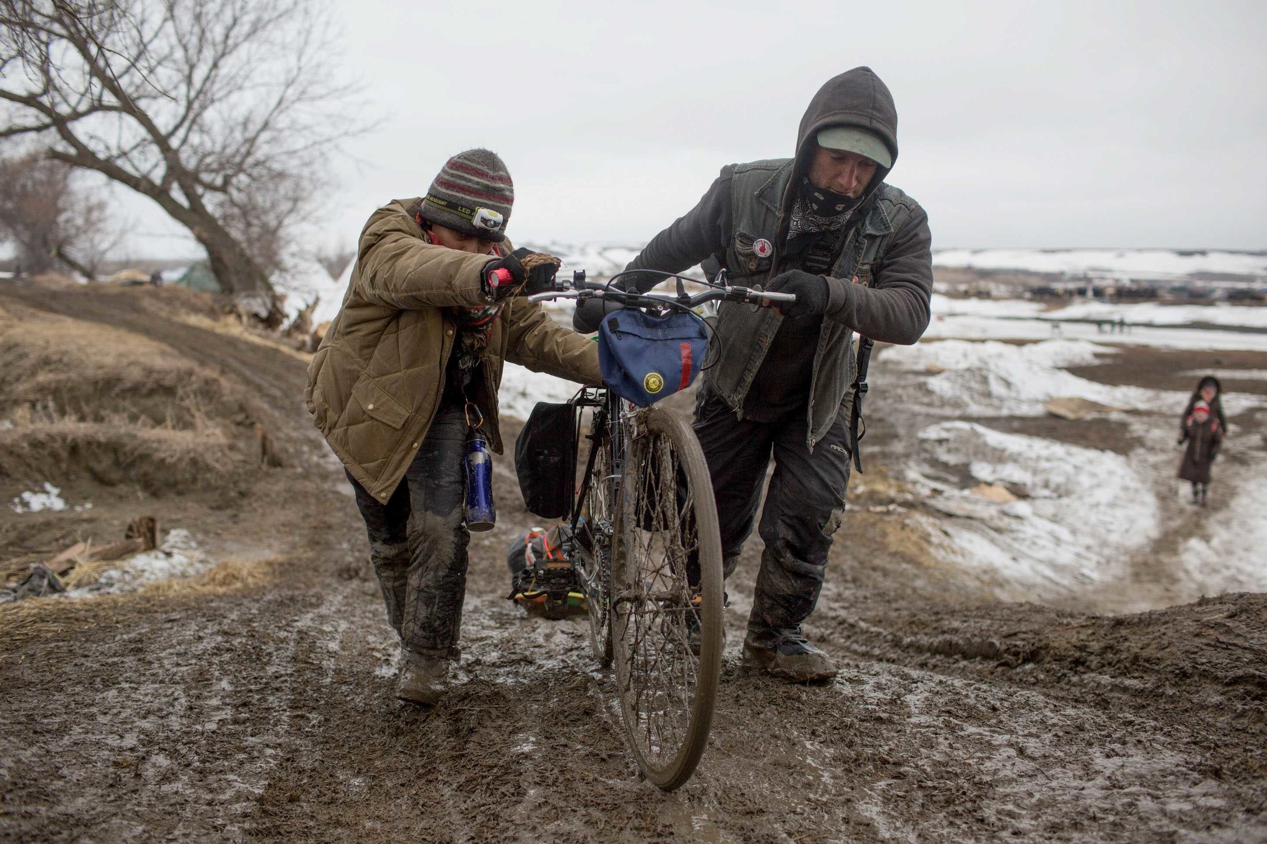  Water protectors push their bike up a hill in an effort to evacuate Rosebud Camp before Federal and state authorities (seen in the distance far right) move over the Cannonball River from Oceti Sakowin into the neighboring protest camps. Rosebud Camp