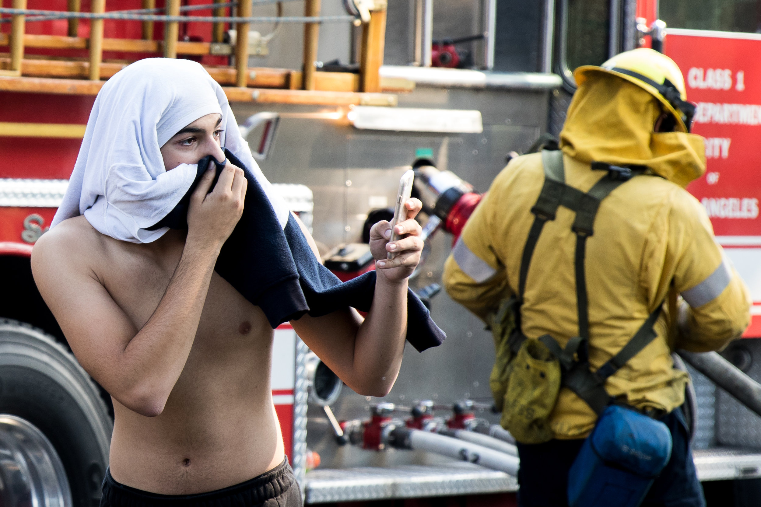  An evacuee of the Skirball Fire records footage of a house burning on his iPhone, while a member of the Los Angeles Fire Department readies a hose to put out the flame. The Skirball Fire which shut down the 405 and burned 450 acres of land on Dec. 6