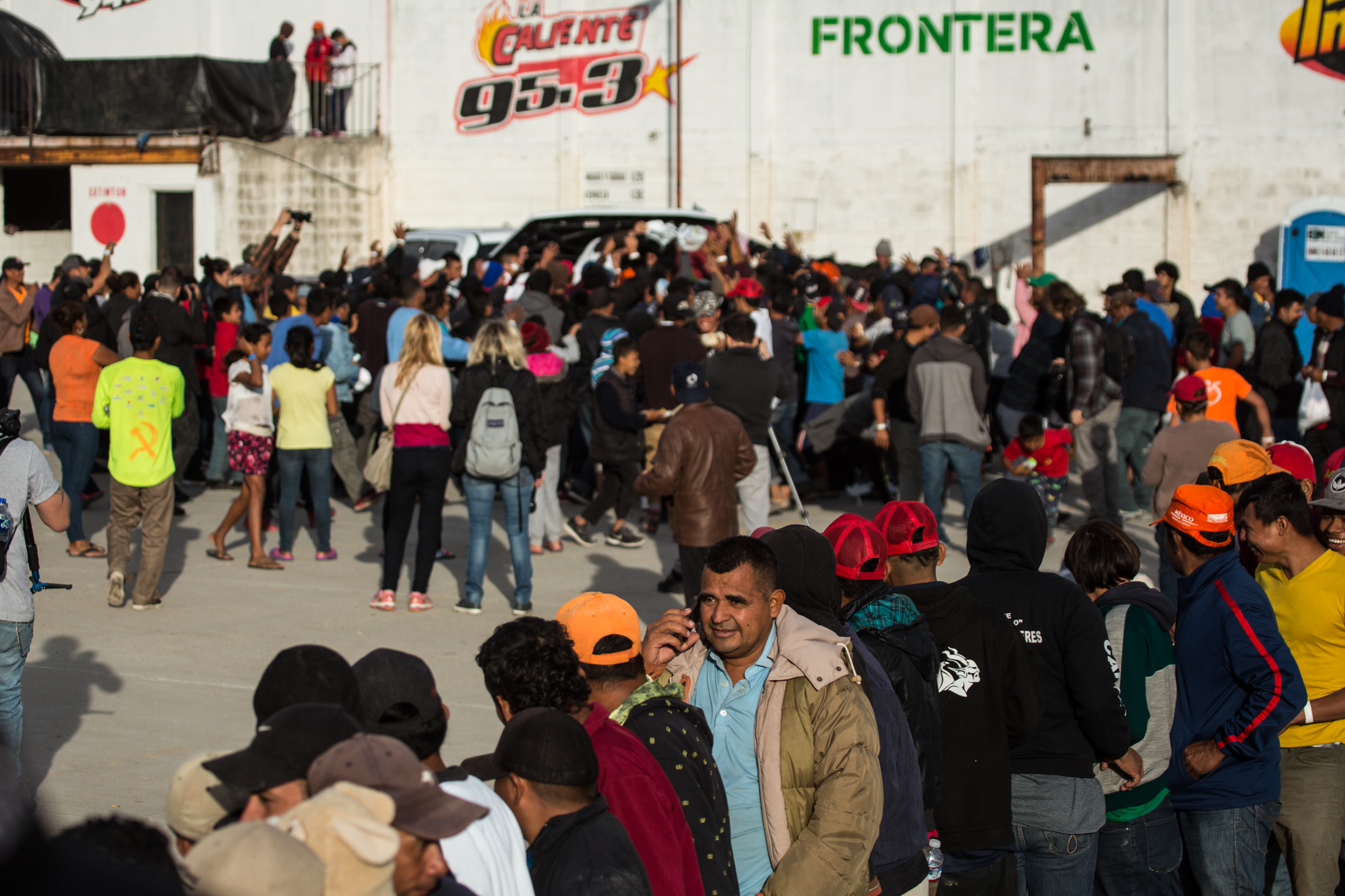   Displaced Hondurans wait in line to receive hot meals prepared by non-government aid groups at El Barretal in Tijuana, Mexico, Saturday, Dec. 1, 2018. In the background, others attempt to catch care packages containing clothing being thrown from a 