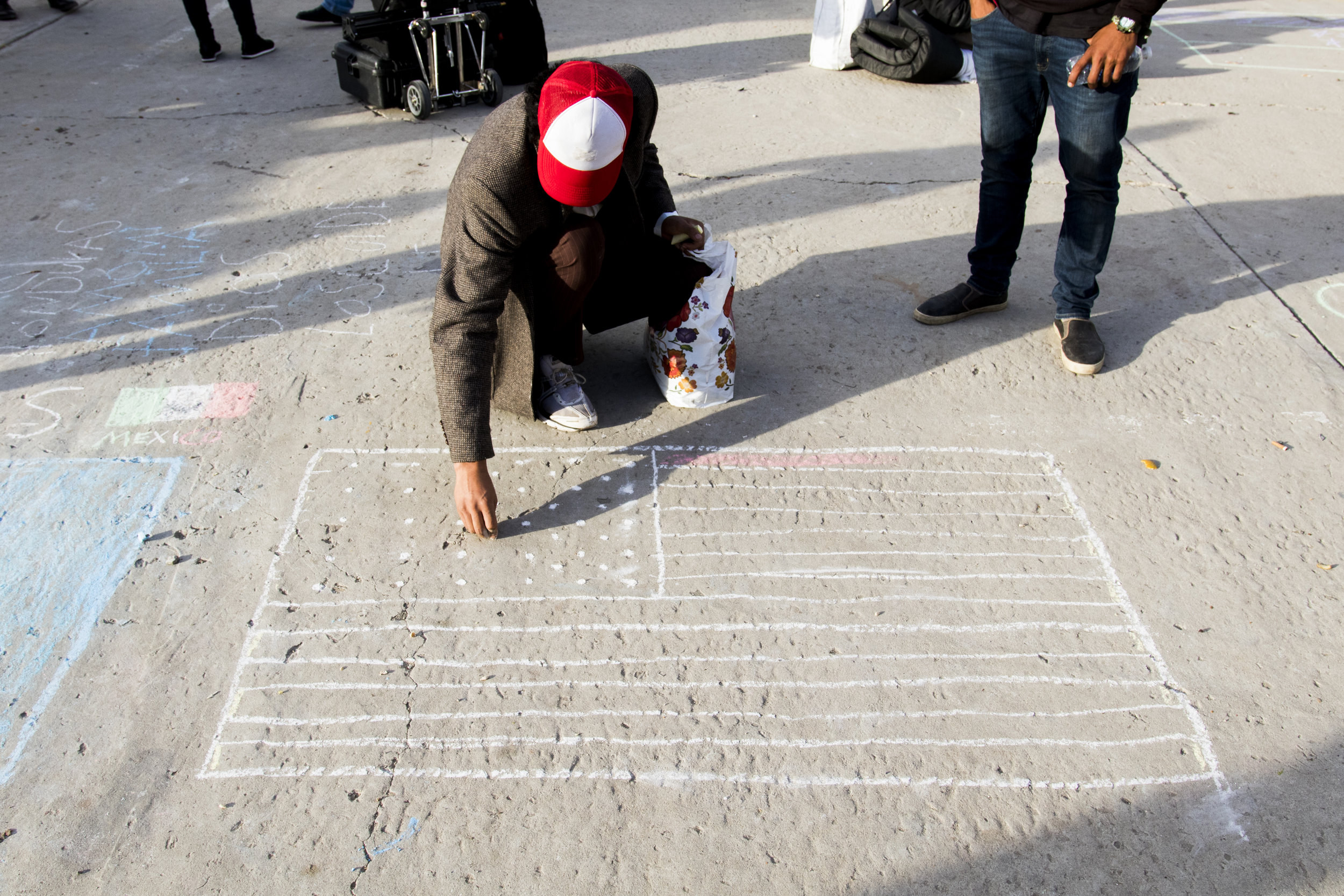  Christian, a migrant male, uses chalk to draw an American Flag on the ground of the El Barretal shelter  in Tijuana, Mexico, Saturday, Dec. 1, 2018. Photo By: Zane Meyer-Thornton/ Corsair Contributor  