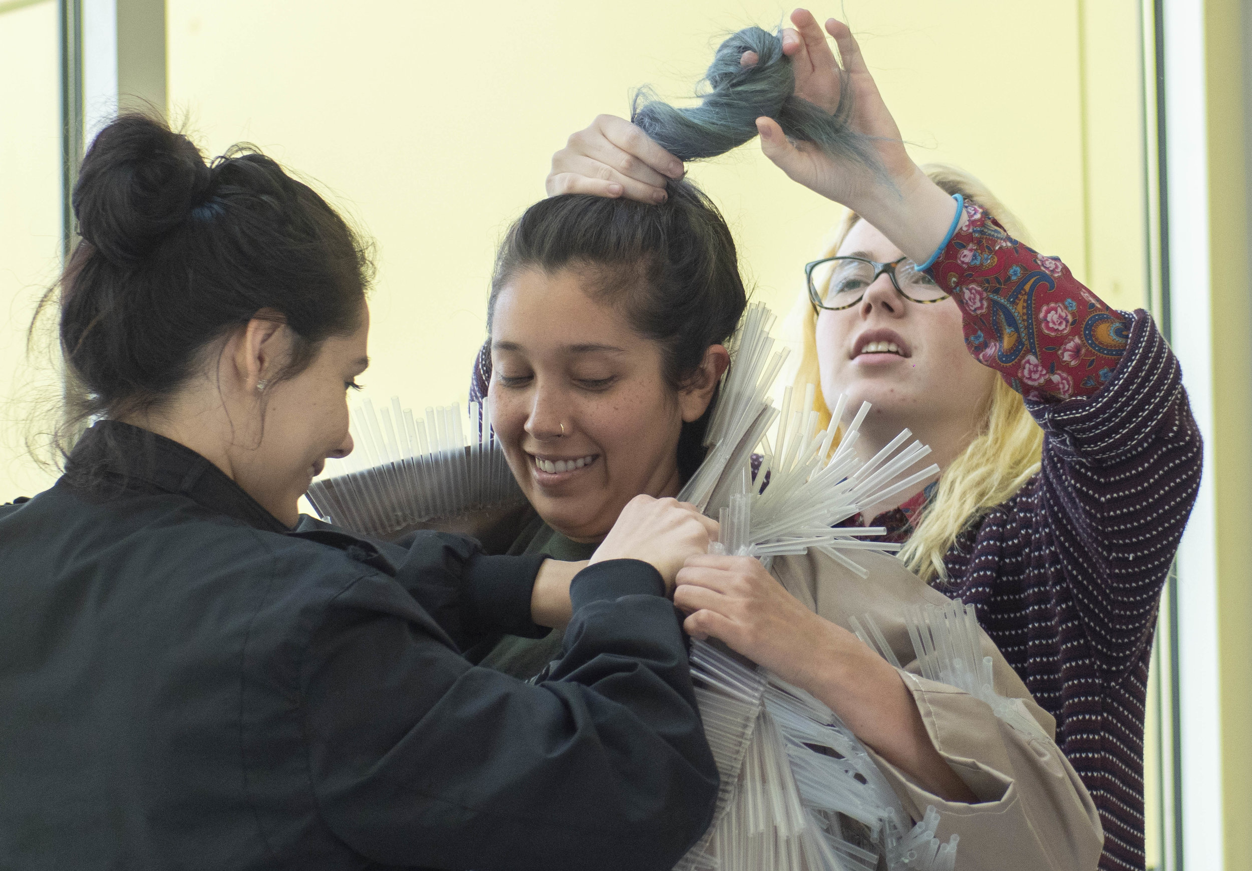  Getting by with a little help from her friends, Carla Claure (center), gets poking straws adjusted away from her cheeks by Lucia Aguilar Cole. Brooke Harrington (right), ties Claure’s hair high and safe from the prickly plastic. She wears the straw 