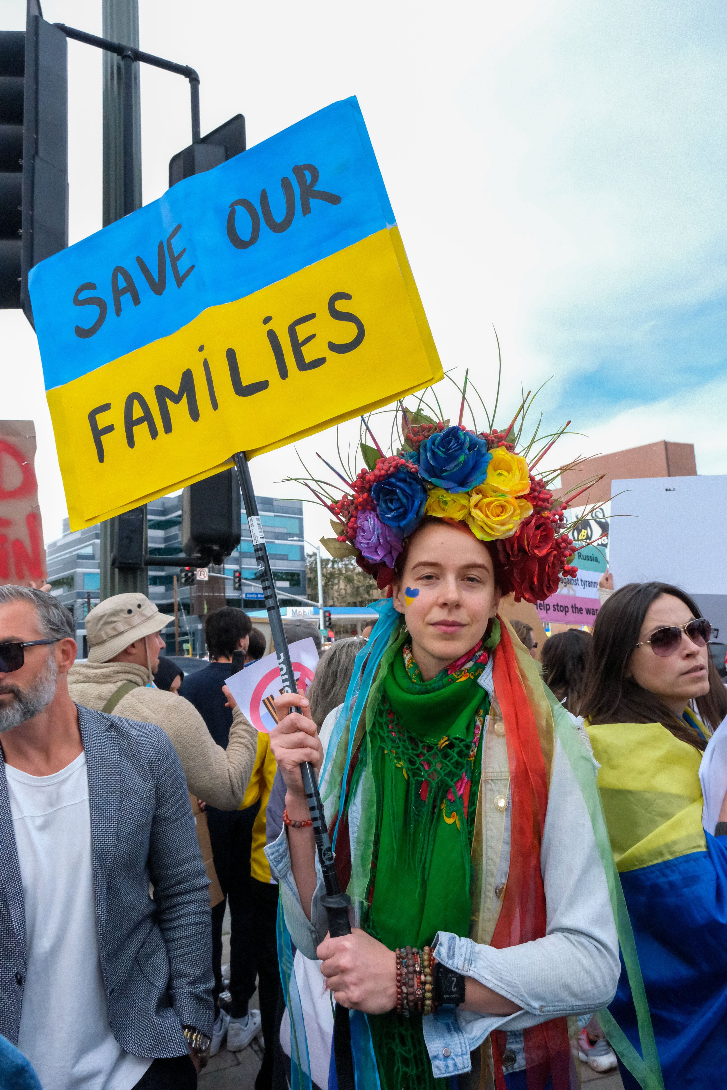  Maryna Volk wears a crown of colored flowers, and holds a sign that says, ‘Save Our Families’. Volk says, “This is heartbreaking. This is surreal, I can’t believe this is Europe, this is my country, Ukraine. No one stands there for us. We are asking