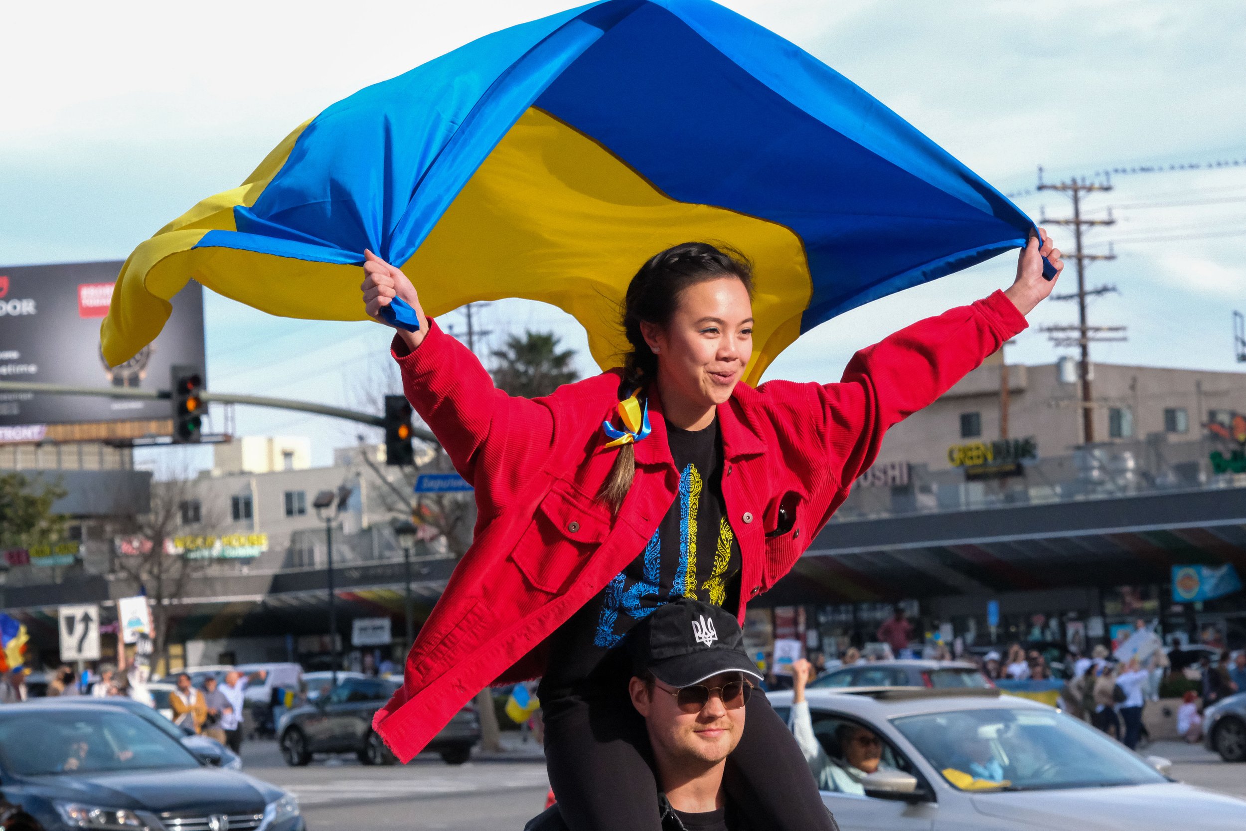  Masha Fam is atop of Oleks Pushkar’s shoulders crossing the street on Sepulveda Boulevard. Fam holds a Ukrainian flag, which blows in the wind as they walk towards the sidewalk. Rally for Ukraine in Westwood, Los Angeles, California on Saturday, Feb