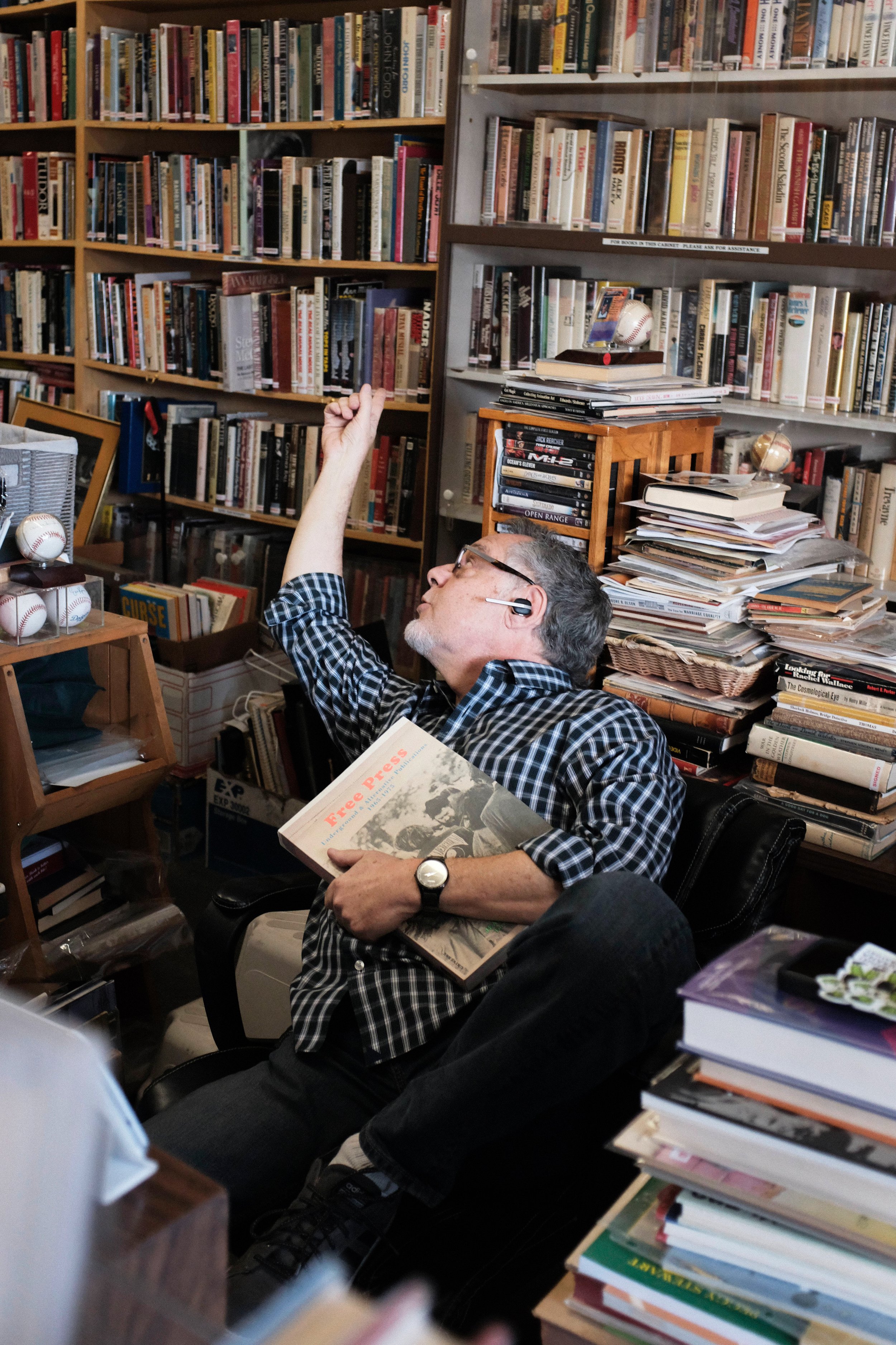  In conversation with a customer who is almost ready to check out their items, David Kaye points out a section that he suggests they should check out. He sits in his chair behind the desk that is surrounded by a vast amount of books. David Kaye Books