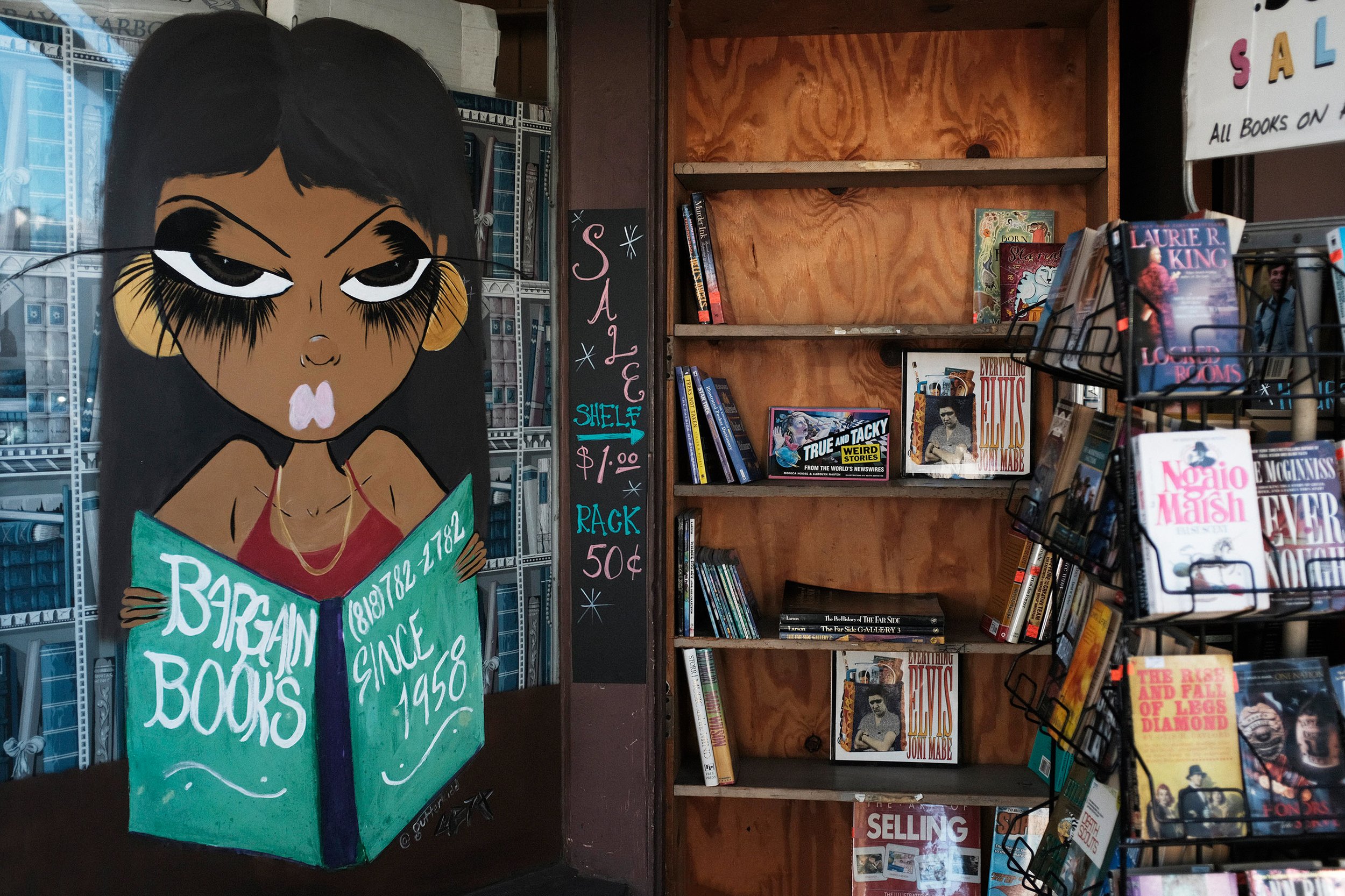  The front of the Bargain Book store features art by the artist written as 'streetkidd,’ of a woman holding a book that shows the shop’s telephone number, and “Since 1958”. There are a few racks of books in the front of the store for .50 and a $1 she