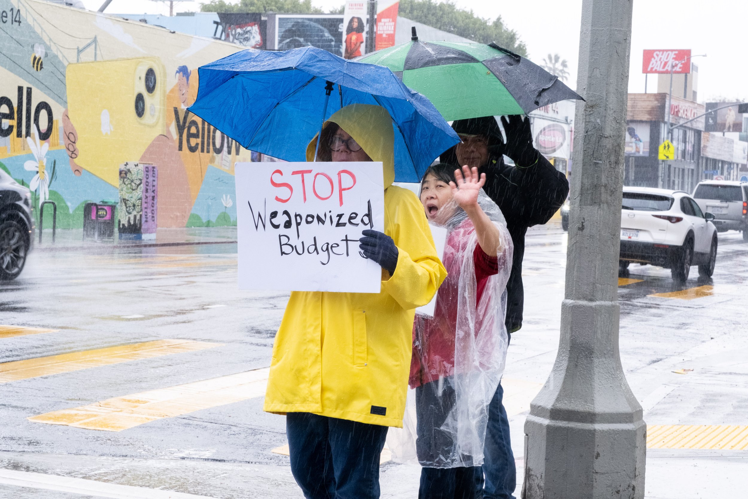  Tracy Cook, wearing a bright yellow rain coat, holds a sign saying "STOP Weaponized Budgets." Her son grew up in the Los Angeles Unified School District, and she continues to be an advocate for public education. She stands with other protestors in f