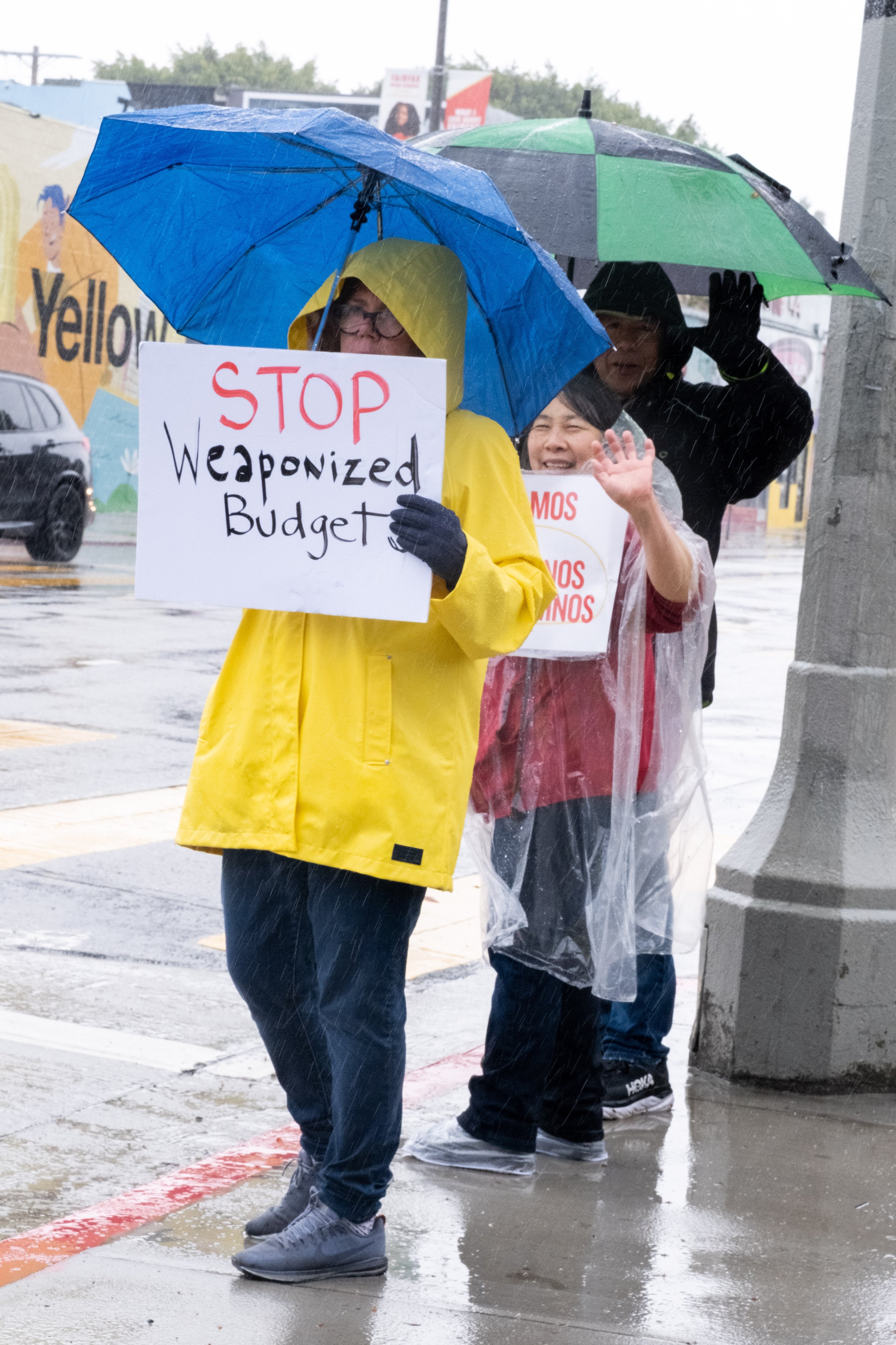  Tracy Cook, wearing a bright yellow rain coat, holds a sign saying "STOP Weaponized Budgets." Her son grew up in the Los Angeles Unified School District, and she continues to be an advocate for public education. She stands with other protestors in f