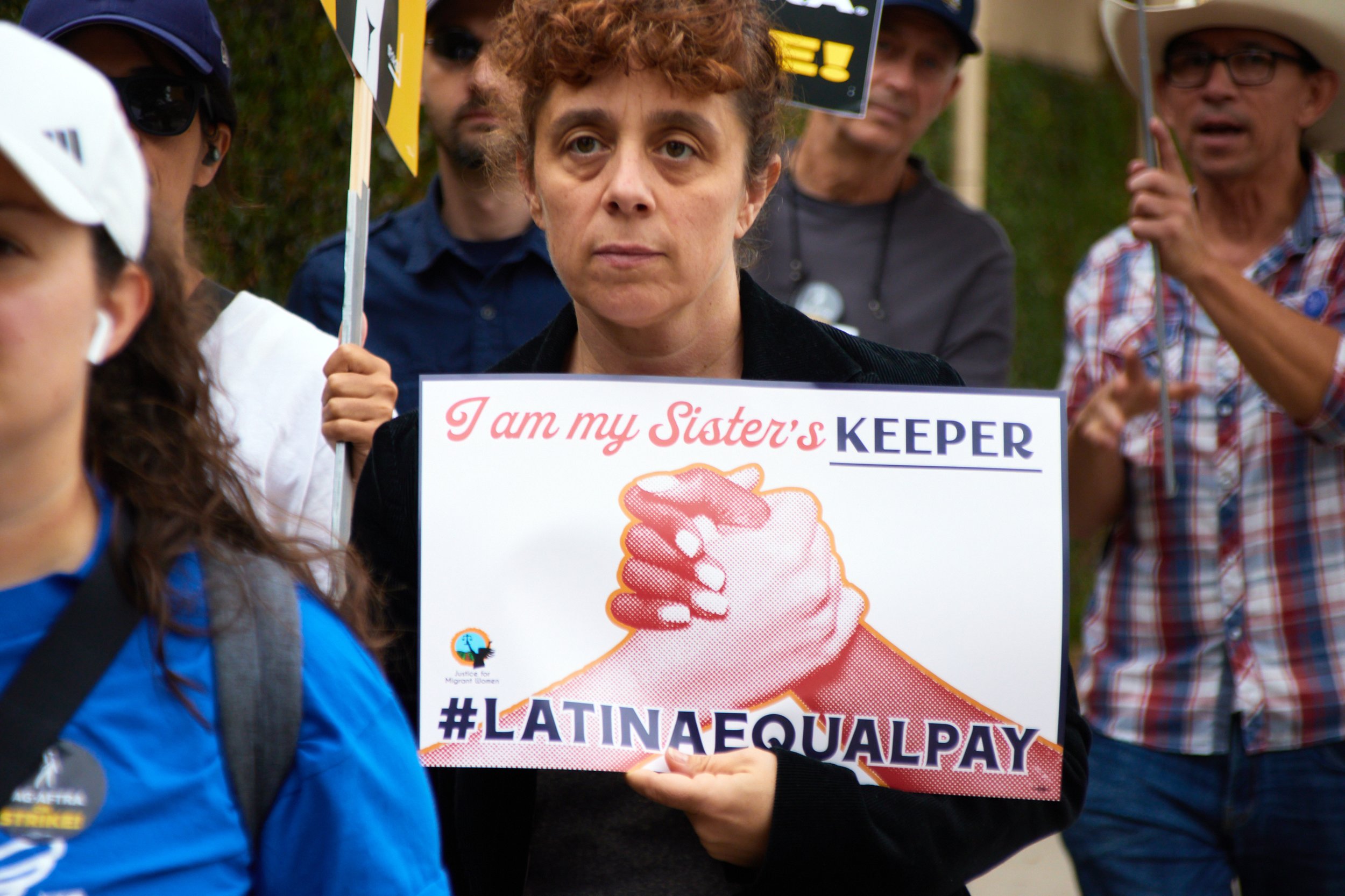  A woman holding a sign with "I am my Sister's KEEPER", "#LATINAEQUALPAY" written on alongside the "Justice for Migrant Women" logo marches alongside picketers during the SAG-AFTRA strike in front of Warner Bros. Studio, Burbank, Calif., Sept 29, 202