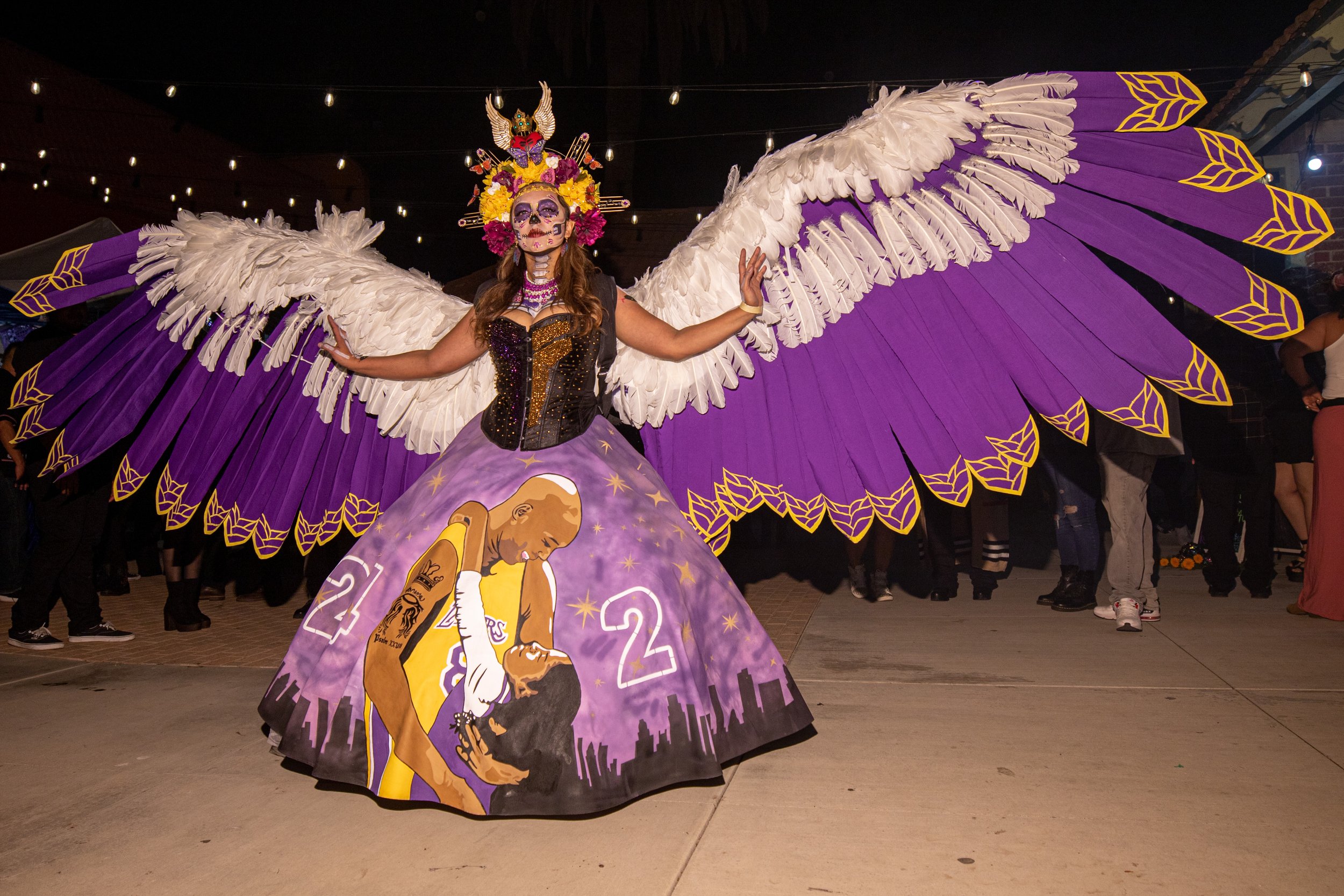  "La Catrina Chicana", wearing a Catrina outfit she designed, stands with her wings fully extended, her dress featuring a mural of Kobe and Gigi Bryant. She designed it in 2020 and is wearing it for the first time tonight at El Velorio's 13th Day of 