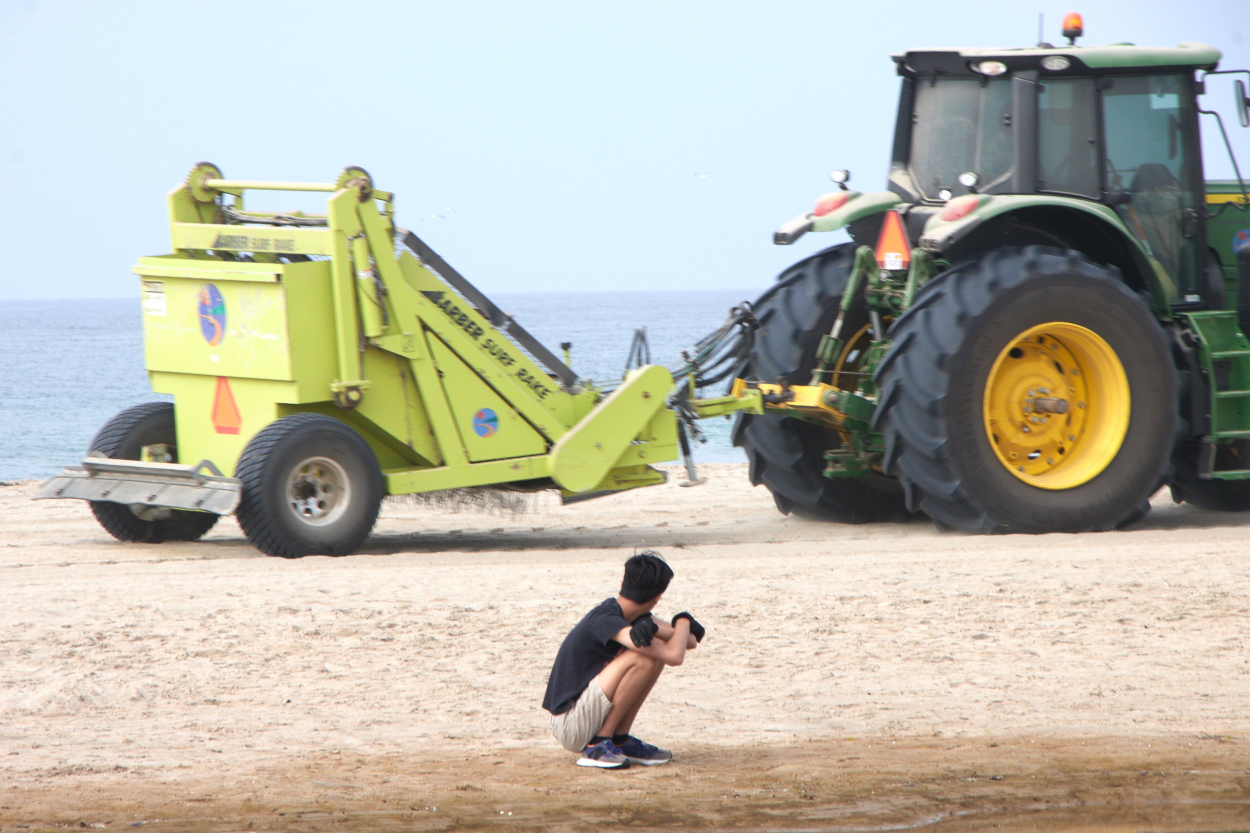  A volunteer squatting watches a tractor towing a beach cleaner drive past during a coastal beach cleanup organized by Santa Monica College Sustainability Center at The Inkwell, Santa Monica, Calif., on Sept 23, 2023. (Danniel Sumarkho | The Corsair)