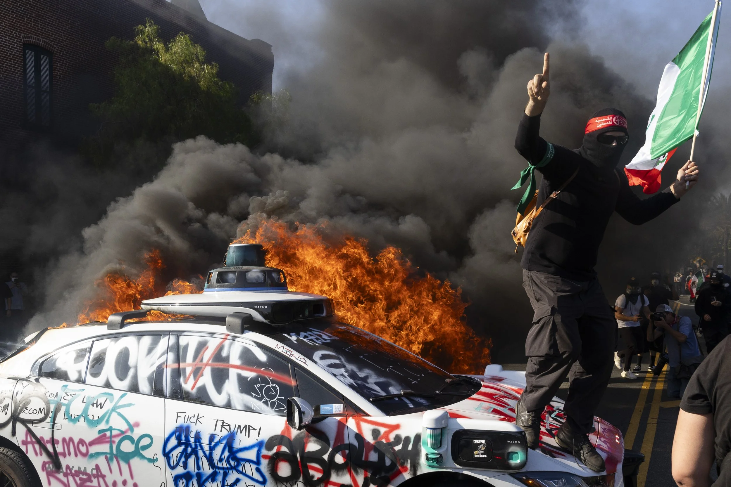 A protester holds a Mexican flag on top of a burning Waymo car while several other Waymo cars are lit on fire, in Downtown Los Angeles, Calif., during the third day of anti-ICE protests on Sunday, June 8, 2025. Protests across Los Angeles County bro