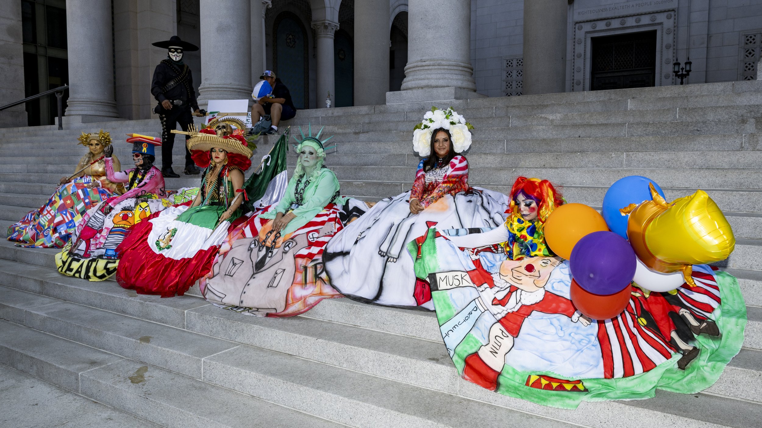 Protesters dressed in colorful dresses sit on the stairs at City Hall in Los Angeles, Calif., on Friday, July 4, 2025. 50501 SOCAL organized a protest called End The Occupation, criticizing the Trump administration’s passing of the Big Beautiful Bil