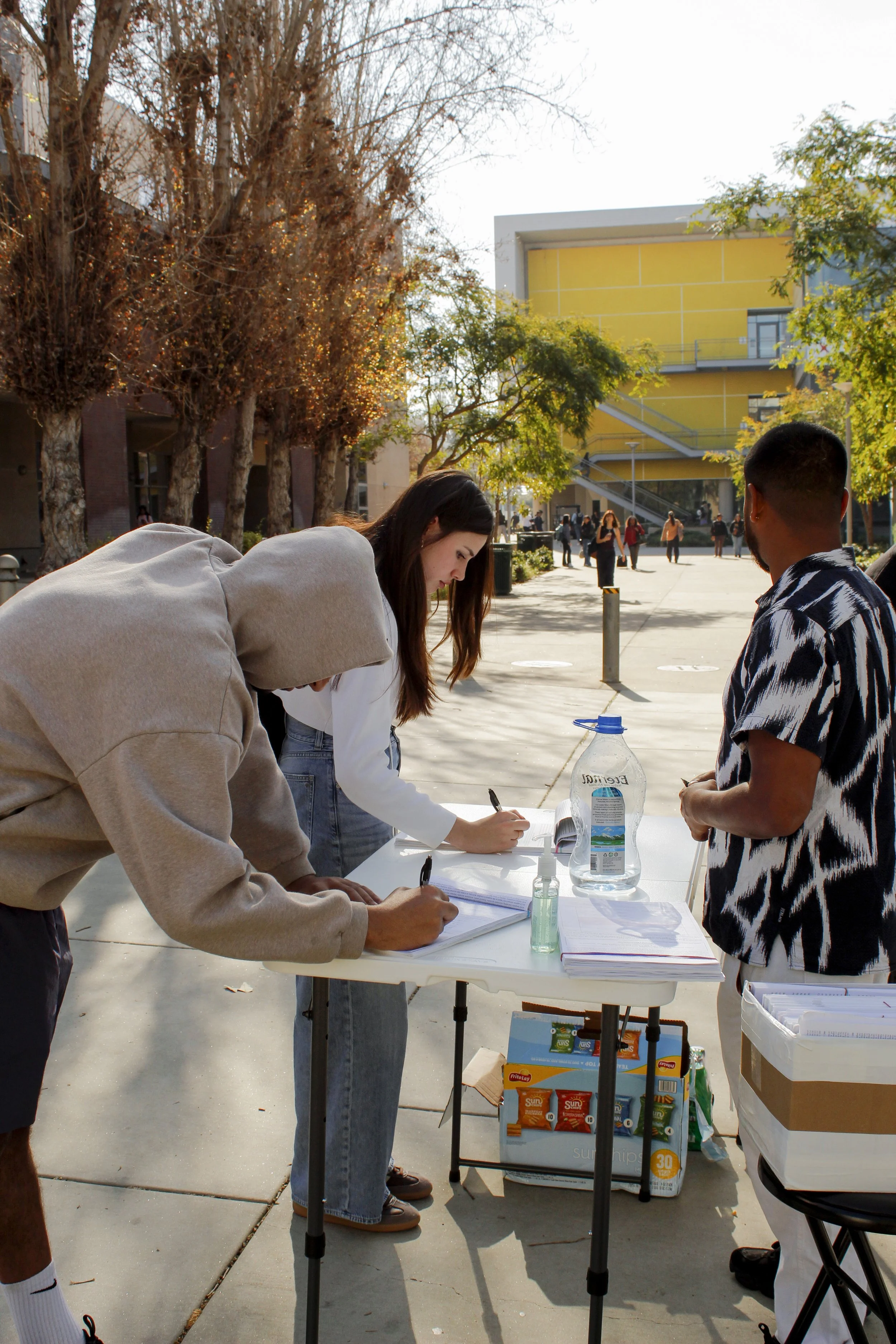  Santa Monica College (SMC) students sign petitions on the SMC main campus on Wednesday, Feb. 25, 2026. Chips and drinks sit in boxes below waiting to be offered to students who agree to sign. Petitioners declined to comment. (Tori Campbell | The Cor