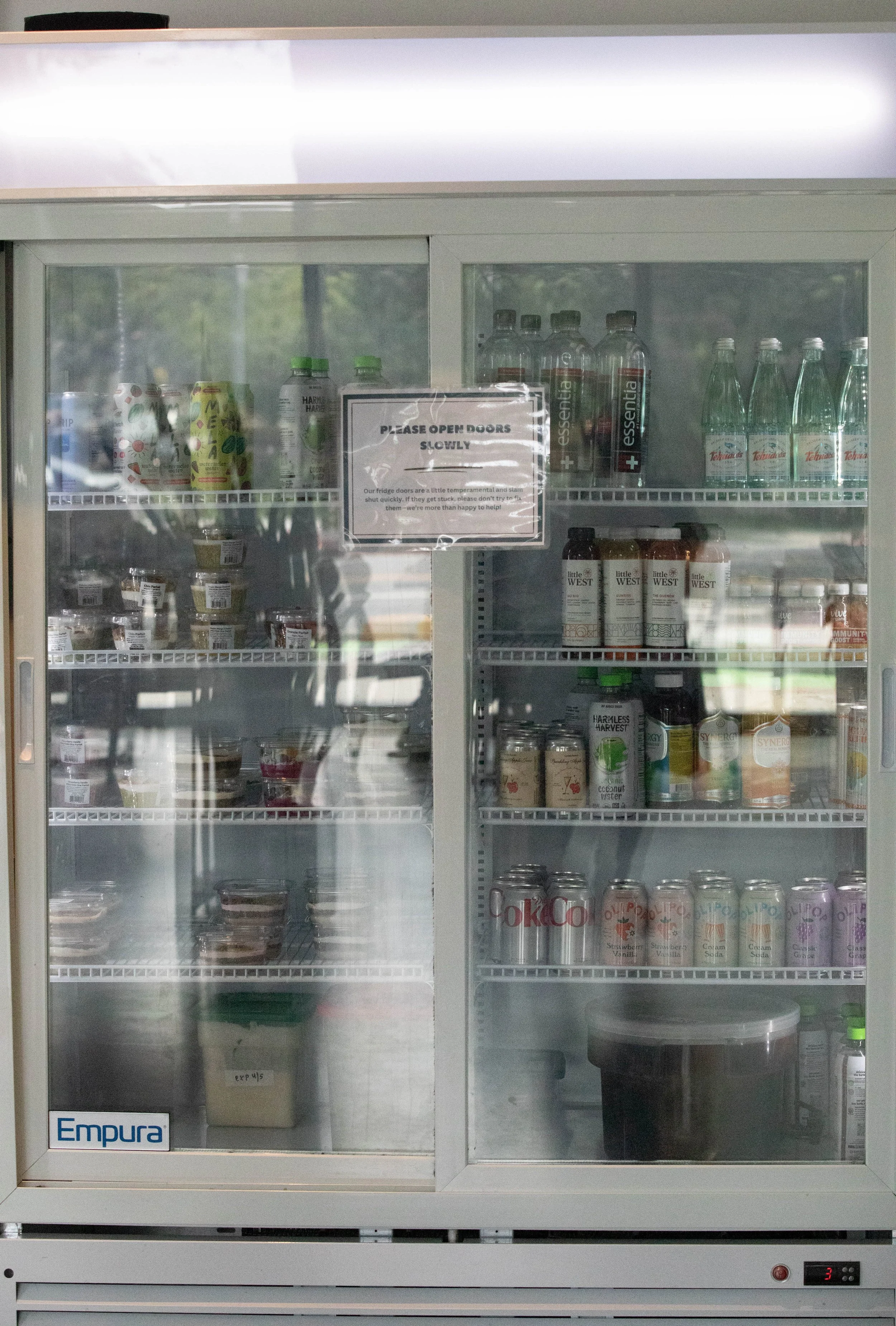  The drink and snack fridge at Cafe Ruisseau contains a number of protein bowls, granola parfaits, and cold bottled drinks. The protein bowls consist of a creamy, fruity base that includes vegan protein and are topped with granola, fruit, and nuts. T