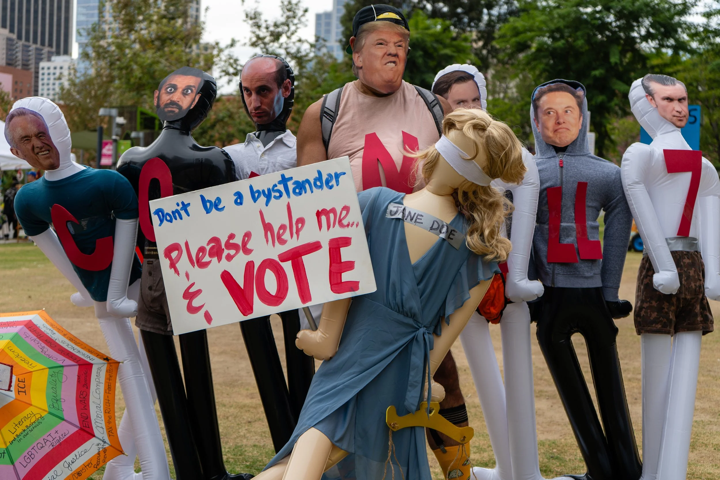  A protestor surrounded by mannequins that have masks of individuals part of the current US administration. A mannequin in the center has a sign that reads 'Don't be a bystander please help me &amp; VOTE' along with a mannequin that has a strip of du