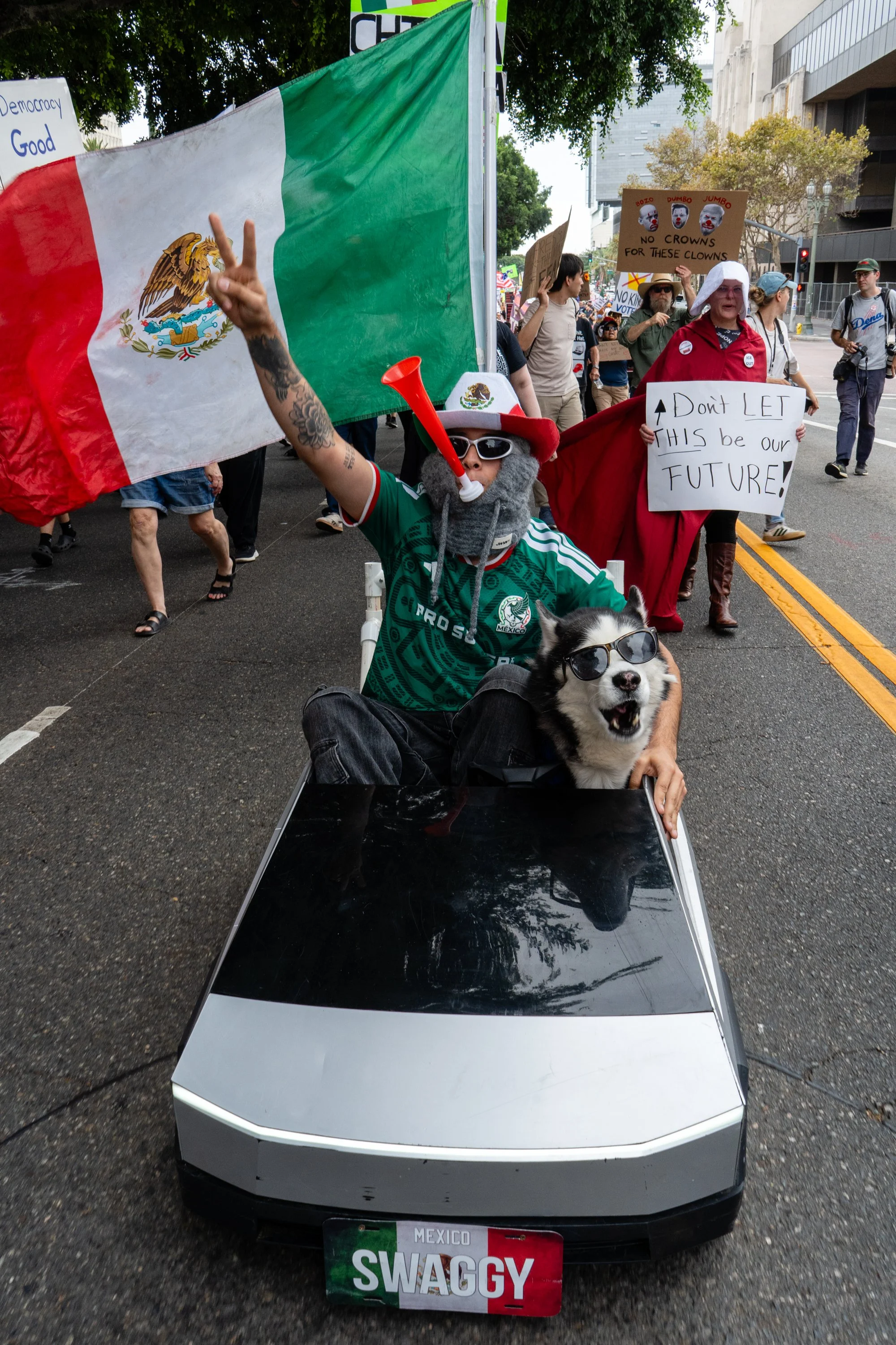  A protestor part of the bigger march wearing a Mexico jersey and sitting in a miniature Tesla Cybertruck with a Mexico flag in the back and a husky as passenger, both donning sunglasses, driving up W. 1st St. on Saturday, Oct. 17, 2026. (Steve Lopez