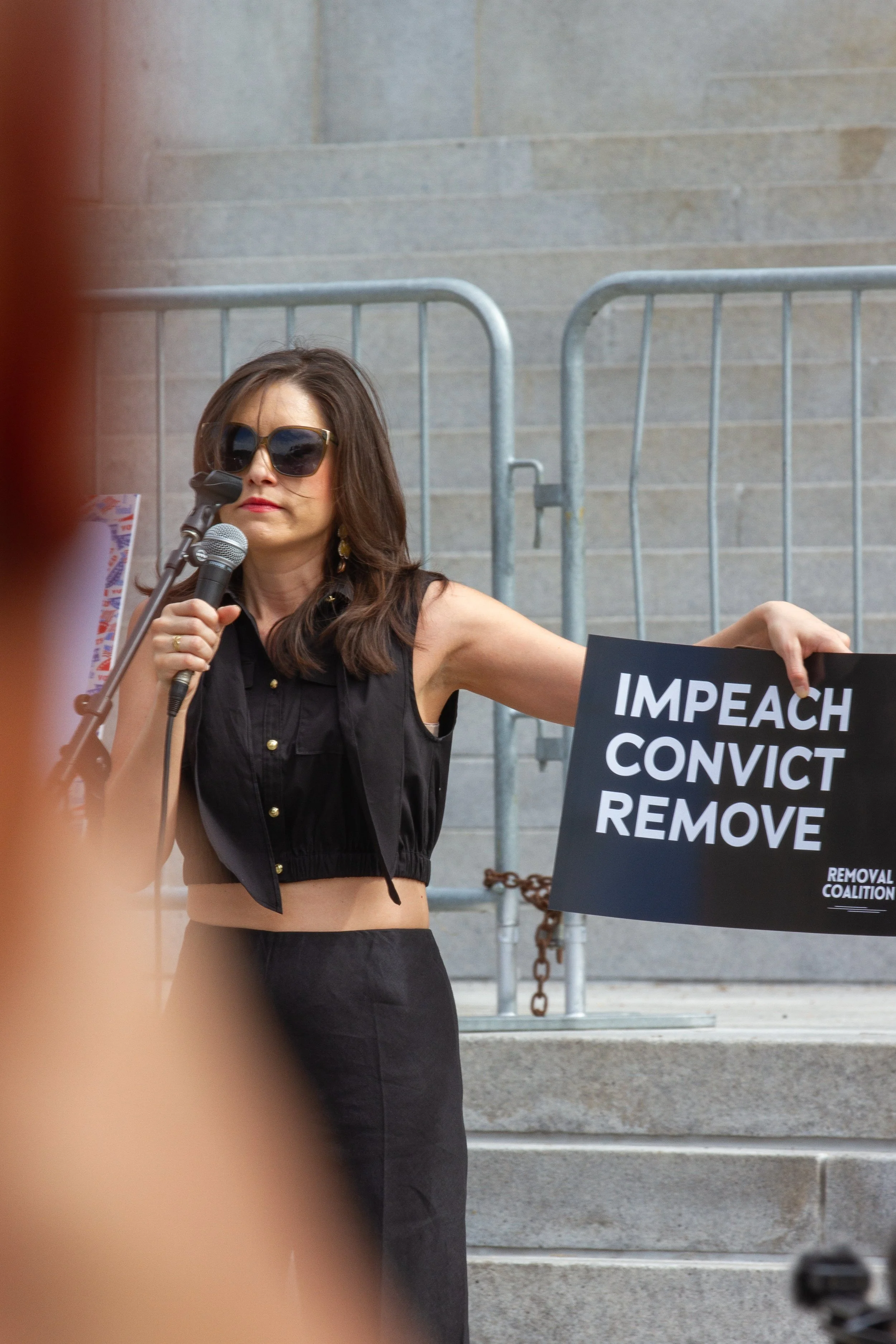  No Kings midterm rally speaker Jessica Denson stands on the front steps of City Hall in downtown LA on Oct. 17, 2026, holding a sign that says “IMPEACH, CONVICT, REMOVE.” Denson worked for President Donald Trump’s 2016 election team, then left after