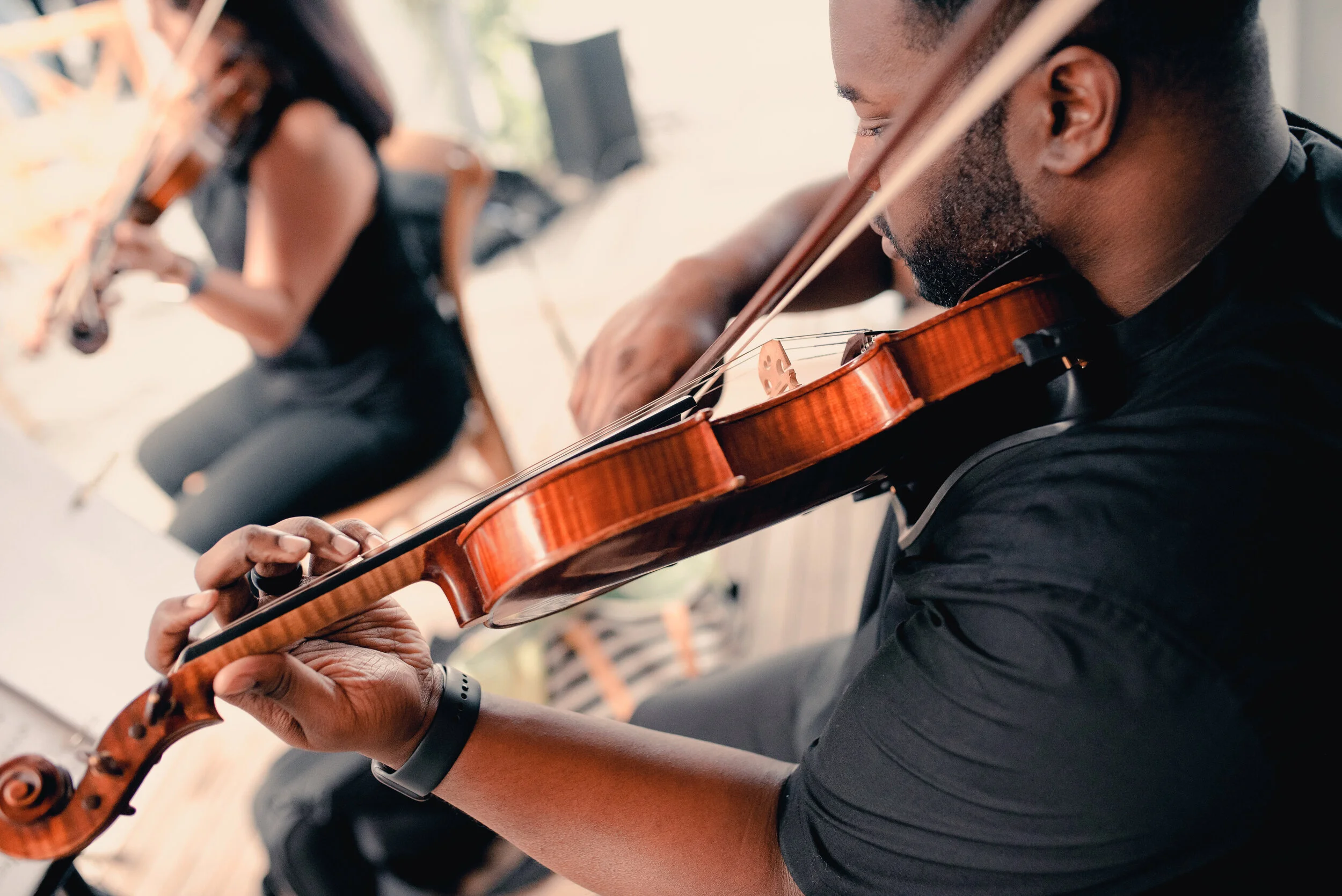 A man playing a violin with a woman playing a violin in the background, both sitting and practicing indoors.