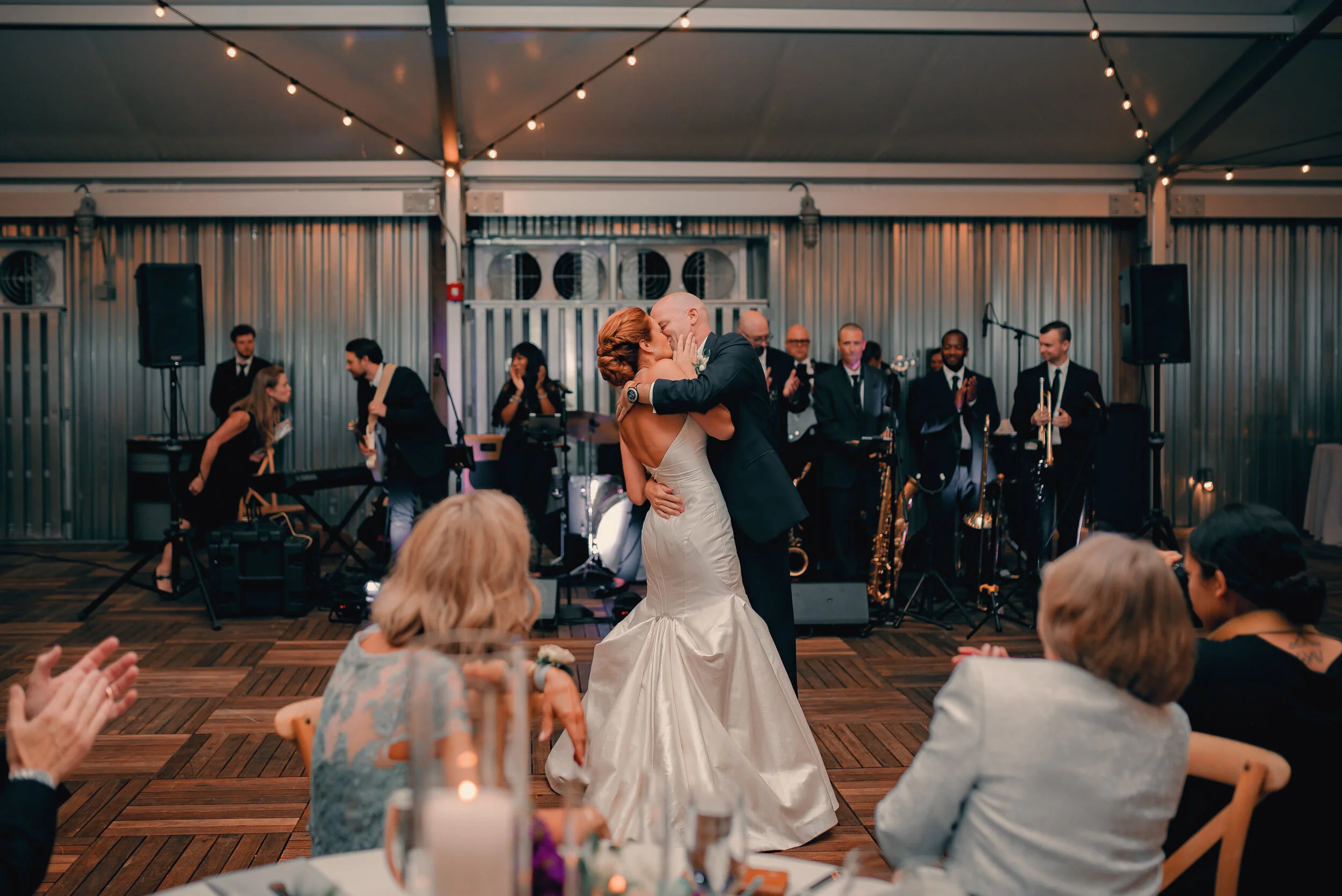 A newlywed couple sharing a romantic dance at their wedding reception, with a band playing in the background and guests clapping and watching.