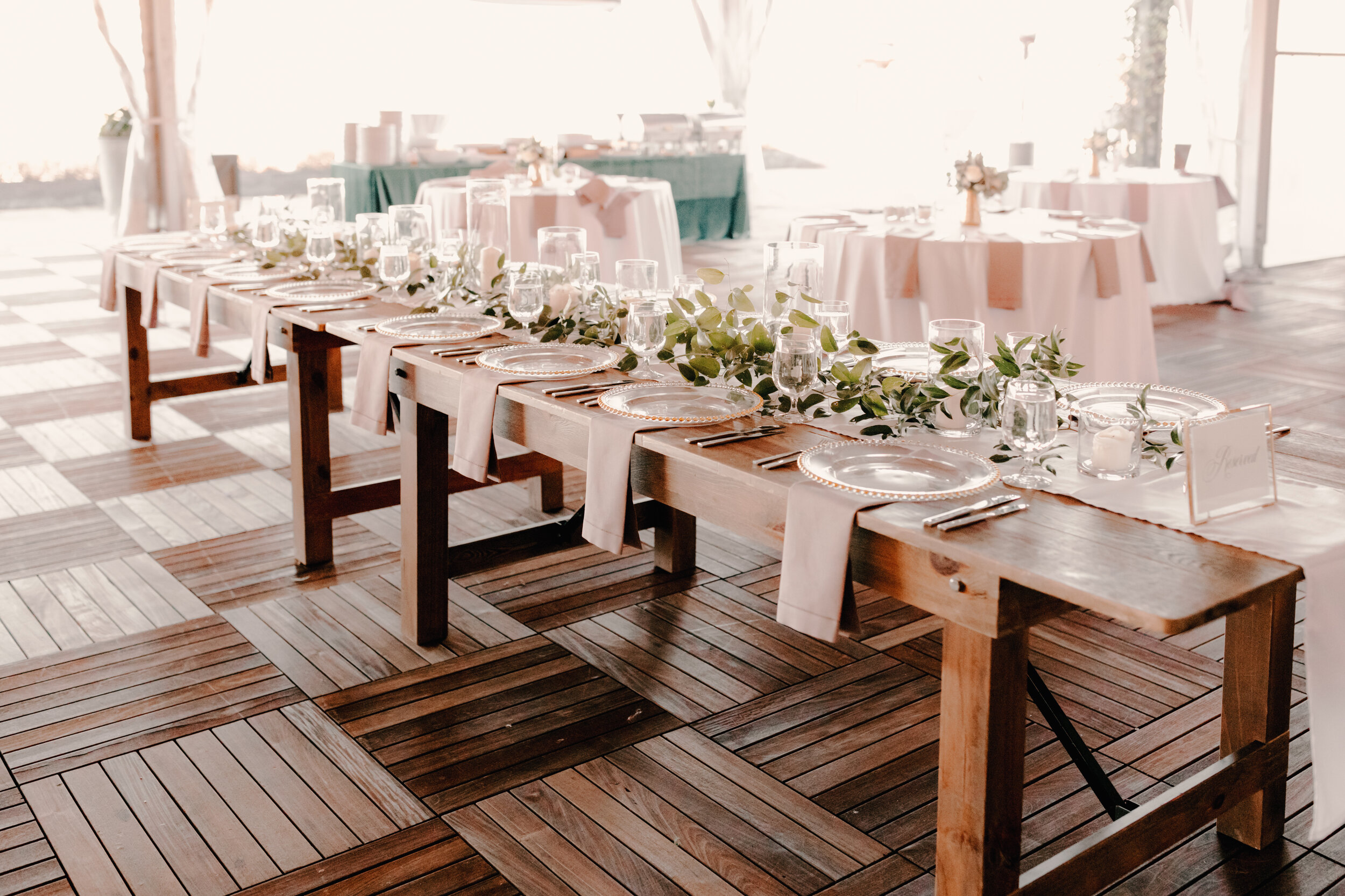 Wedding reception table with place settings, glassware, and green leafy decoration on a wooden table in a bright, sunny room.